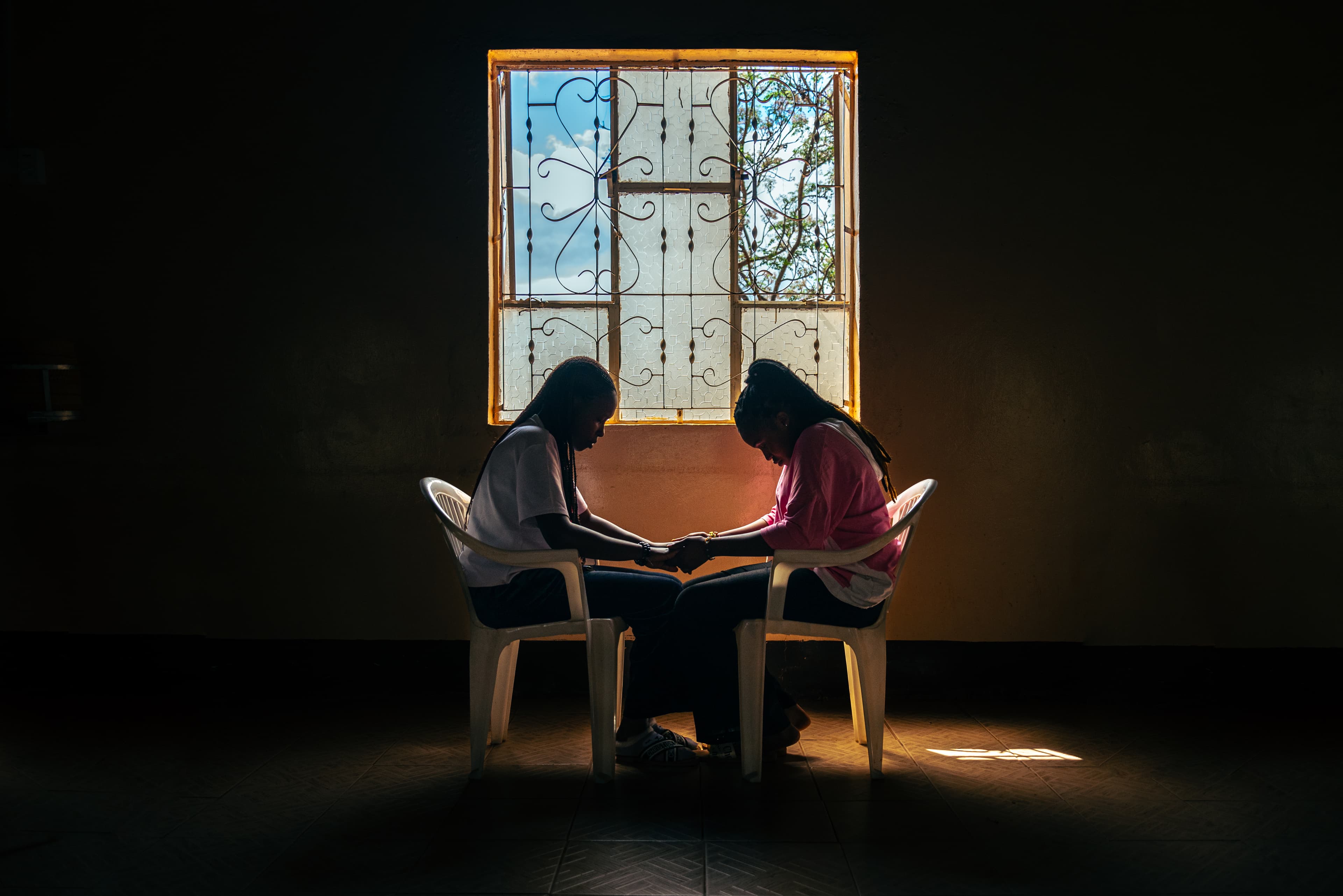 An older woman and a teen girl hold hands in front of a window and pray.