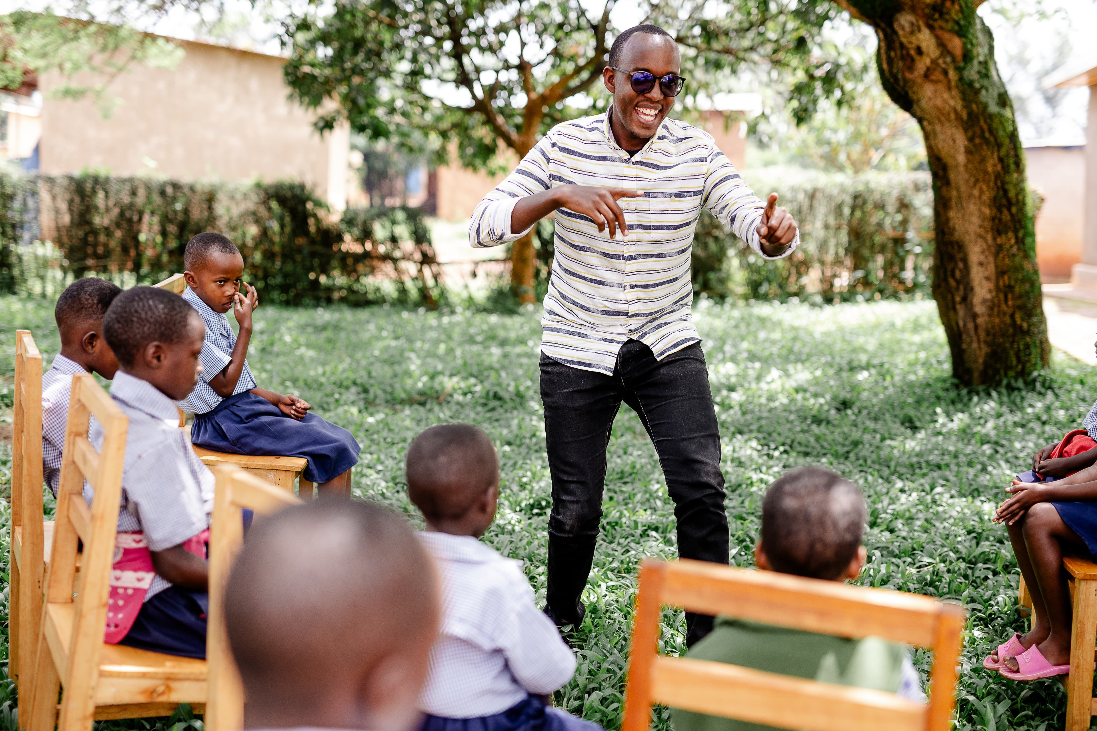 A young African man stands in front of a group of children while smiling.