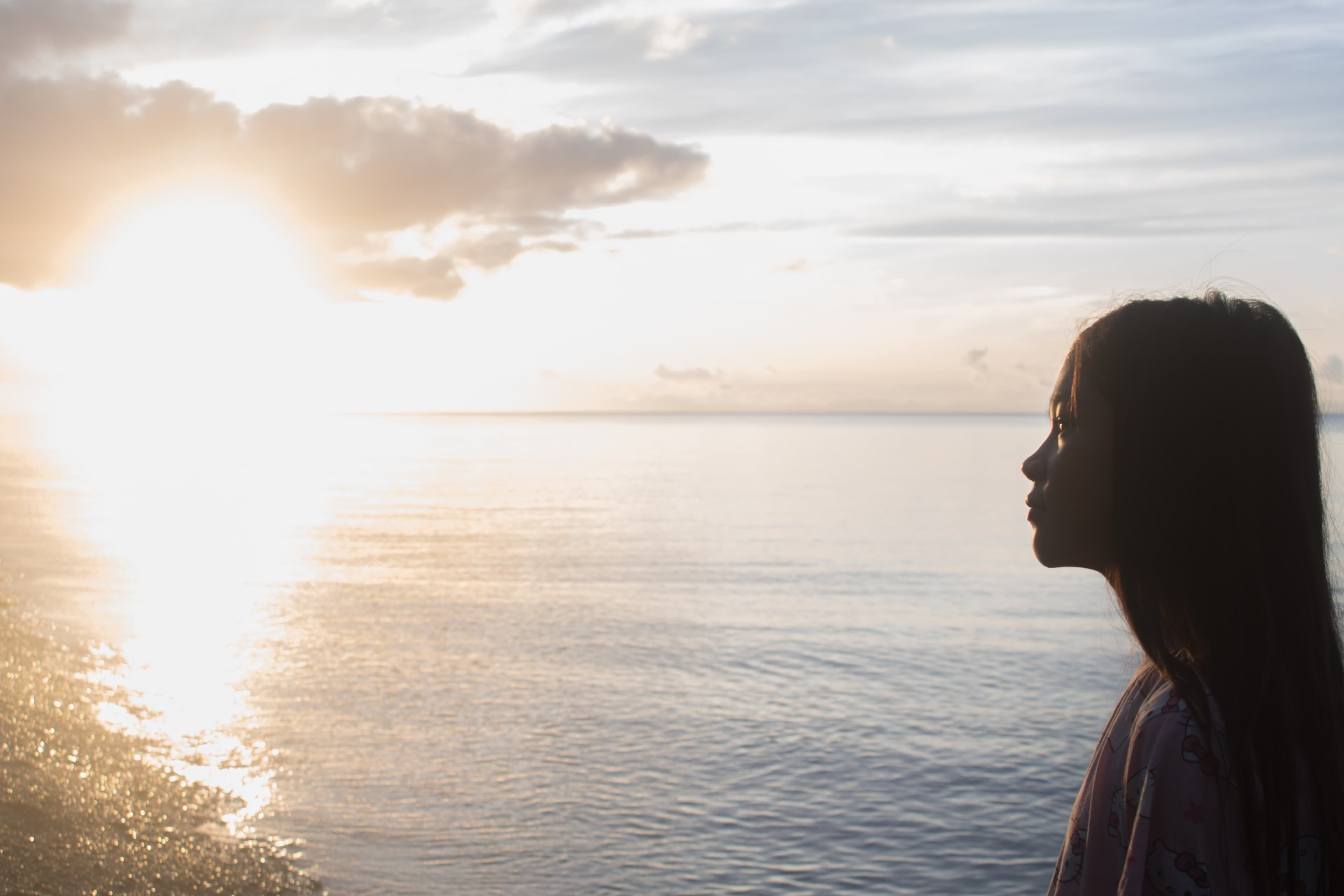 A teen girl looks out across the ocean with the sun shining in from the left.