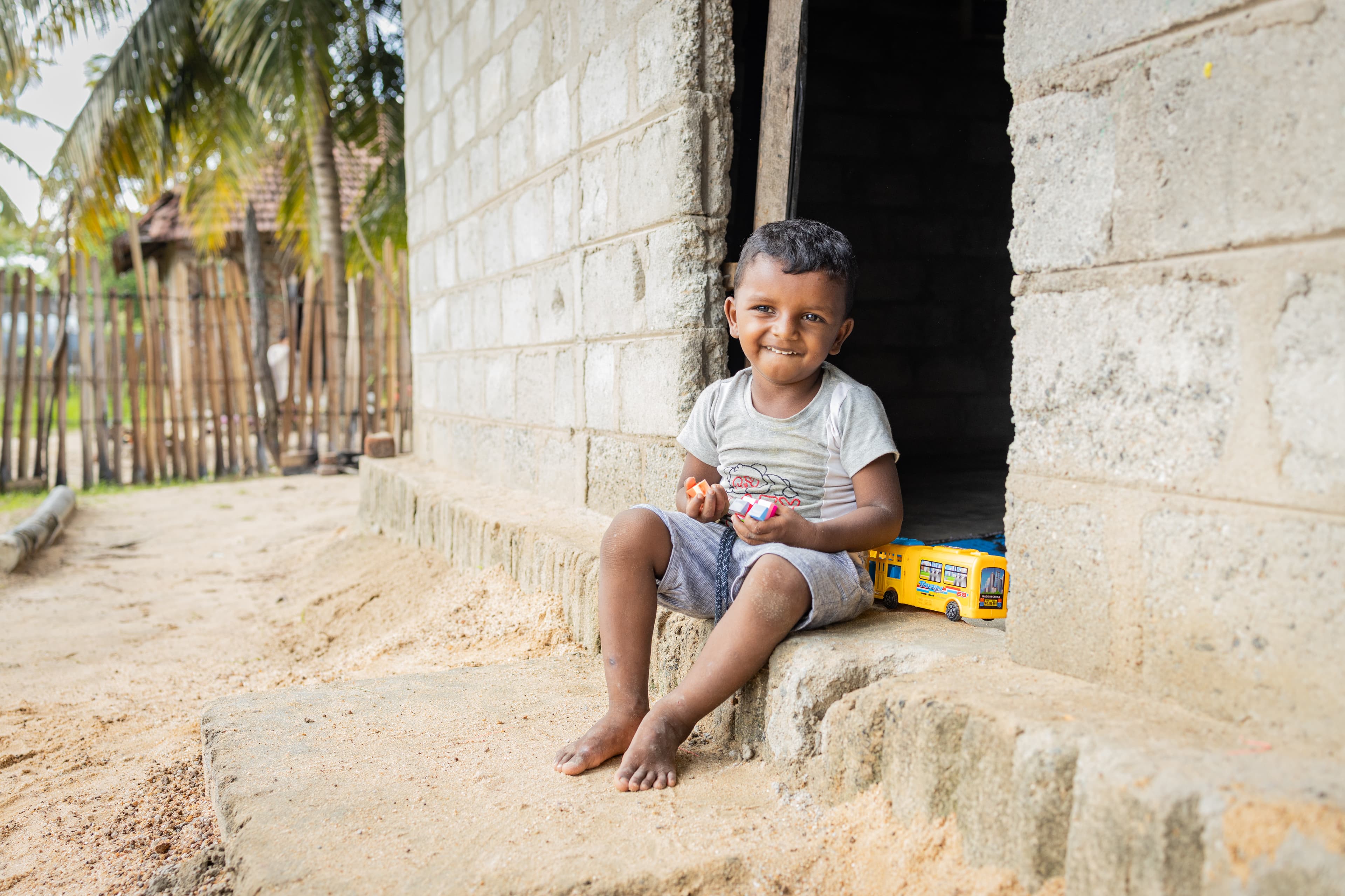 A toddler boy sits barefoot, playing with toys in the doorway of a concrete home. He is wearing a light green T-shirt and blue shorts.