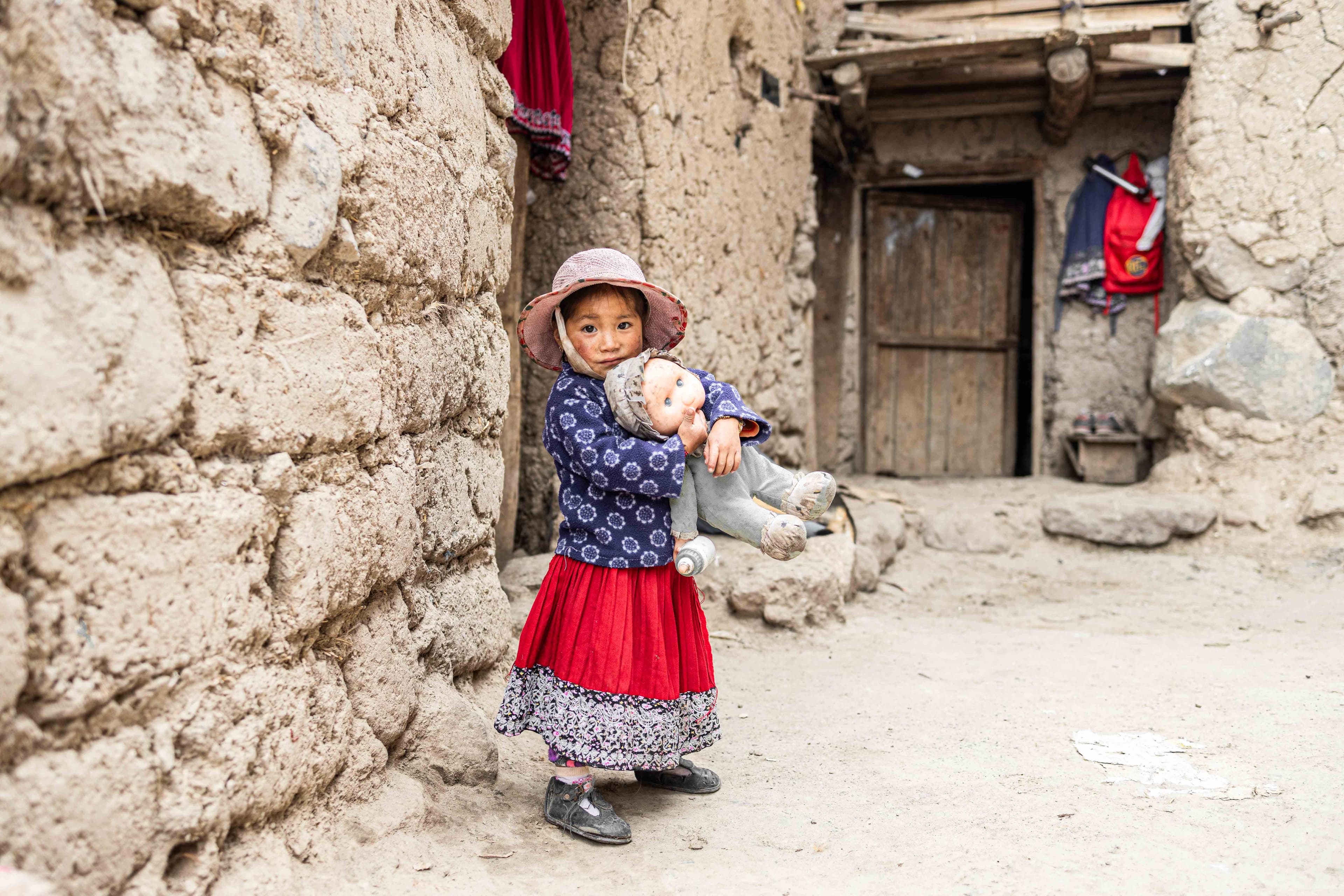 A young Ecuadorean girl holds a doll and stands outside her home.