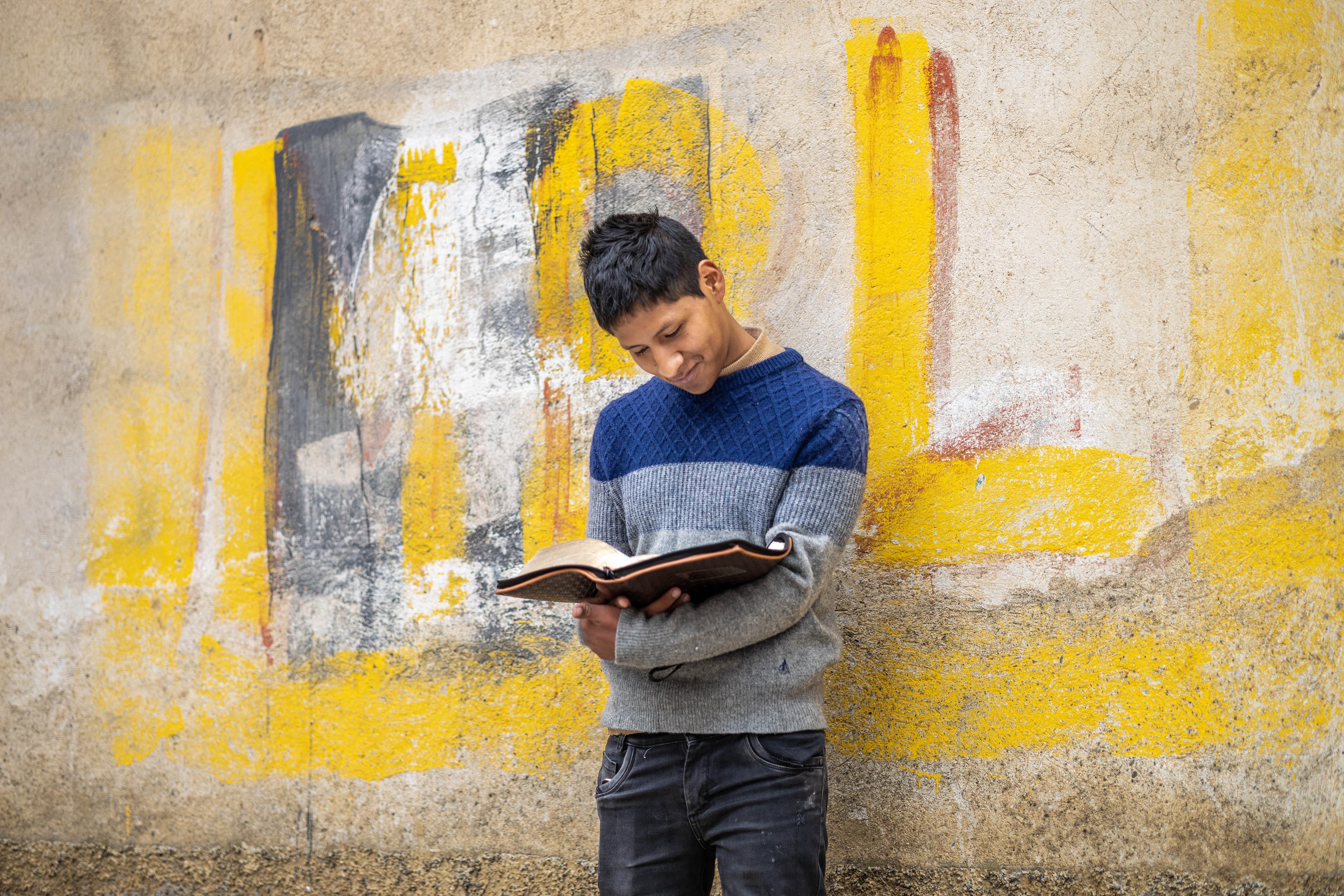 A teen Bolivian boy stands against a wall while holding a reading a Bible.