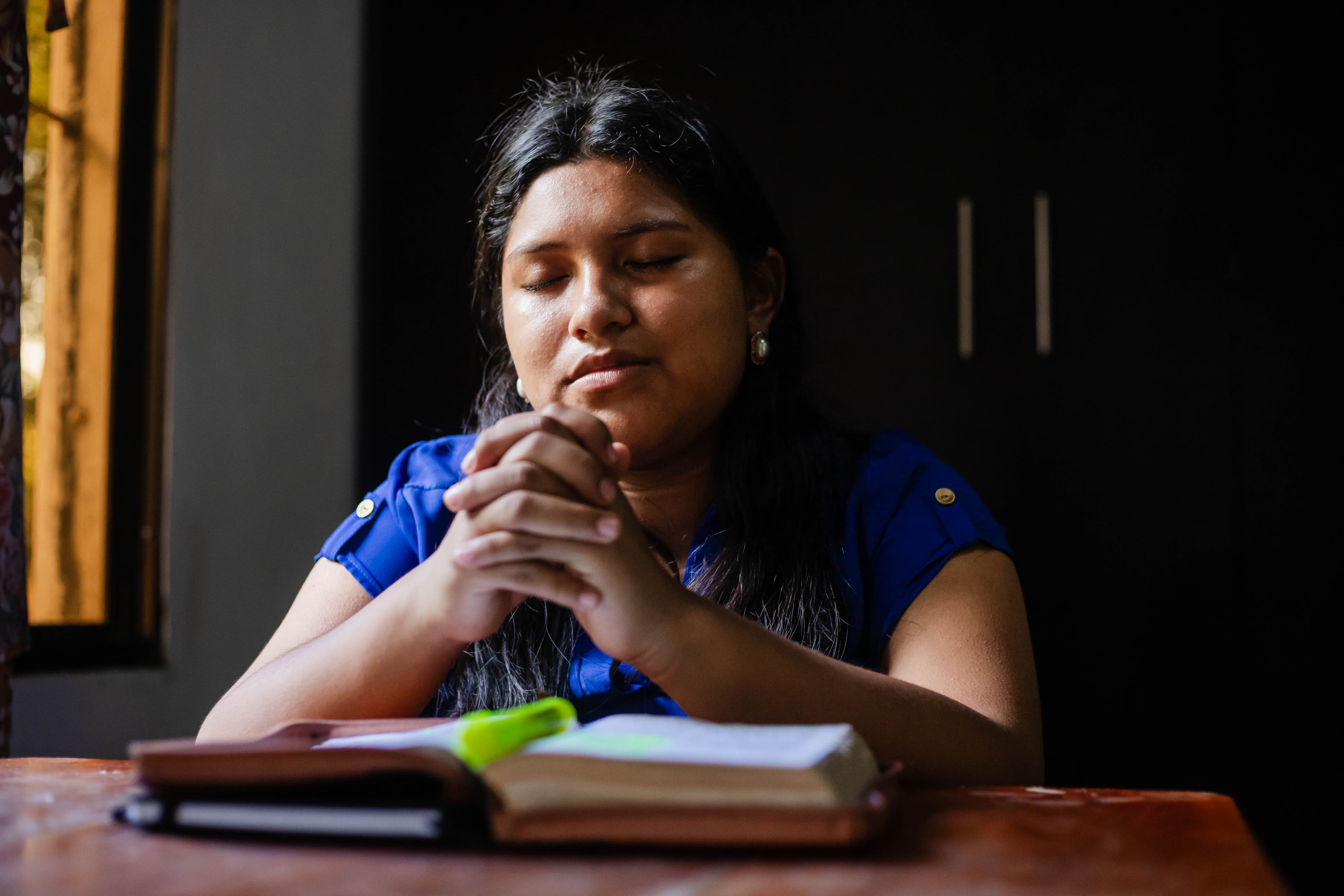 A teen girl sits at a desk with her hands clasped in prayer behind a Bible.