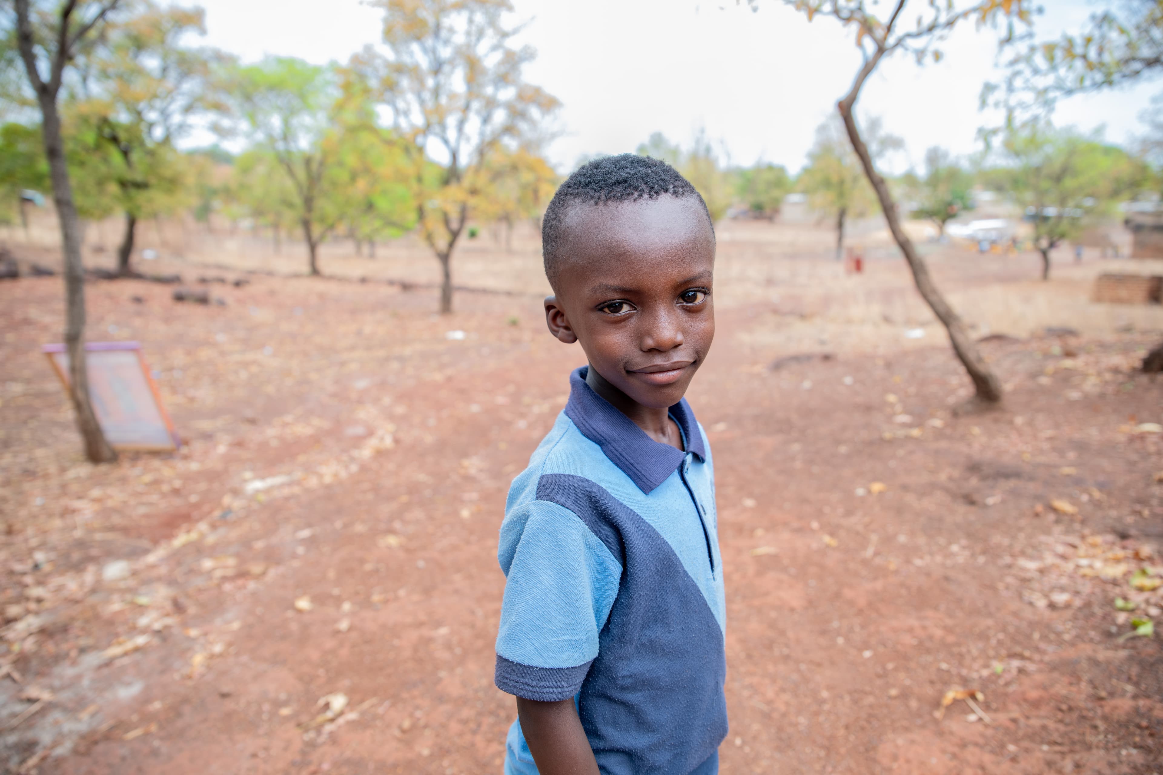 A young boy in a blue polo shirt stands outside in his community, smiling, with trees in the background.