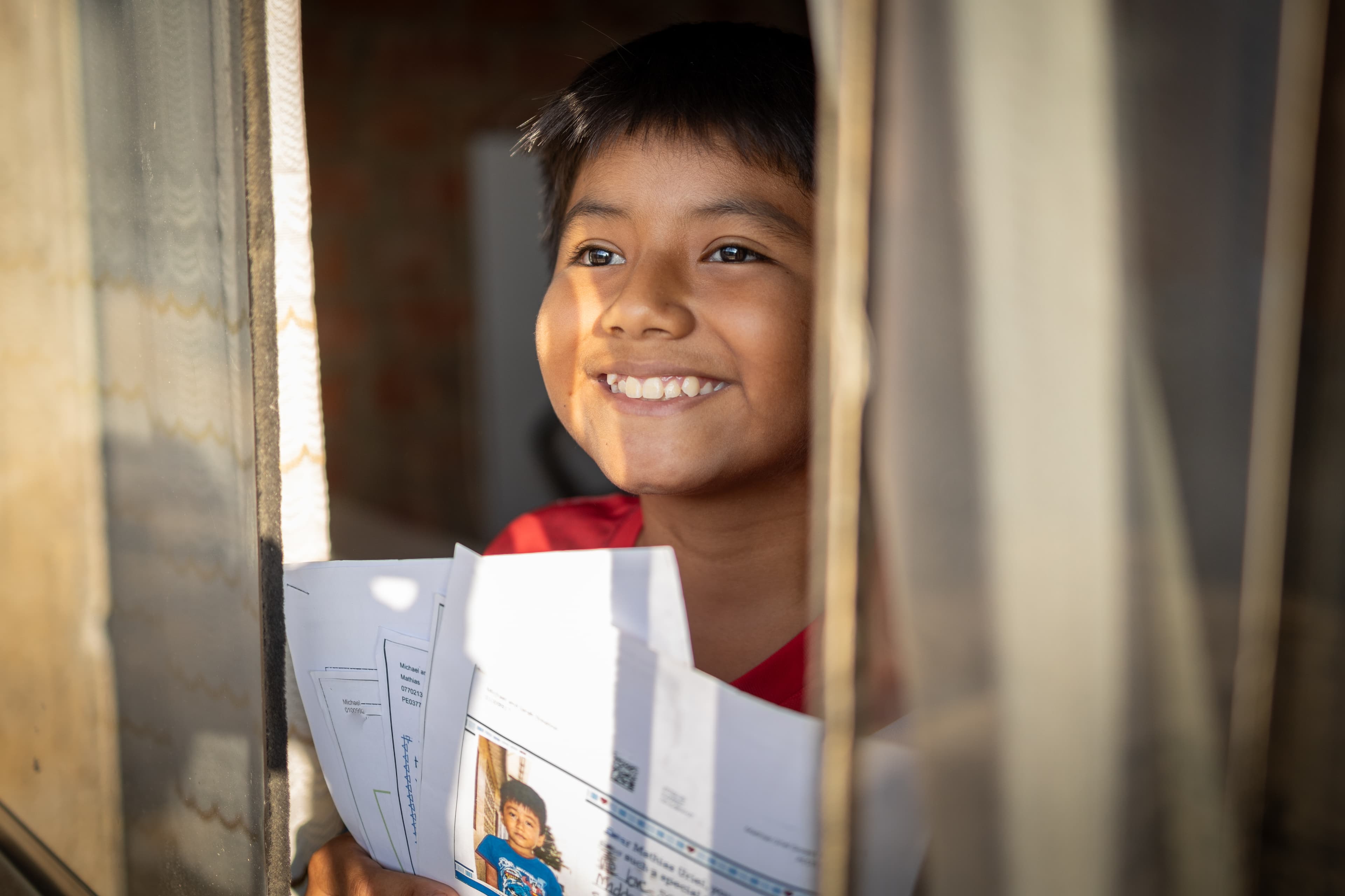 A young boy smiles out a window while holding sponsor letters.