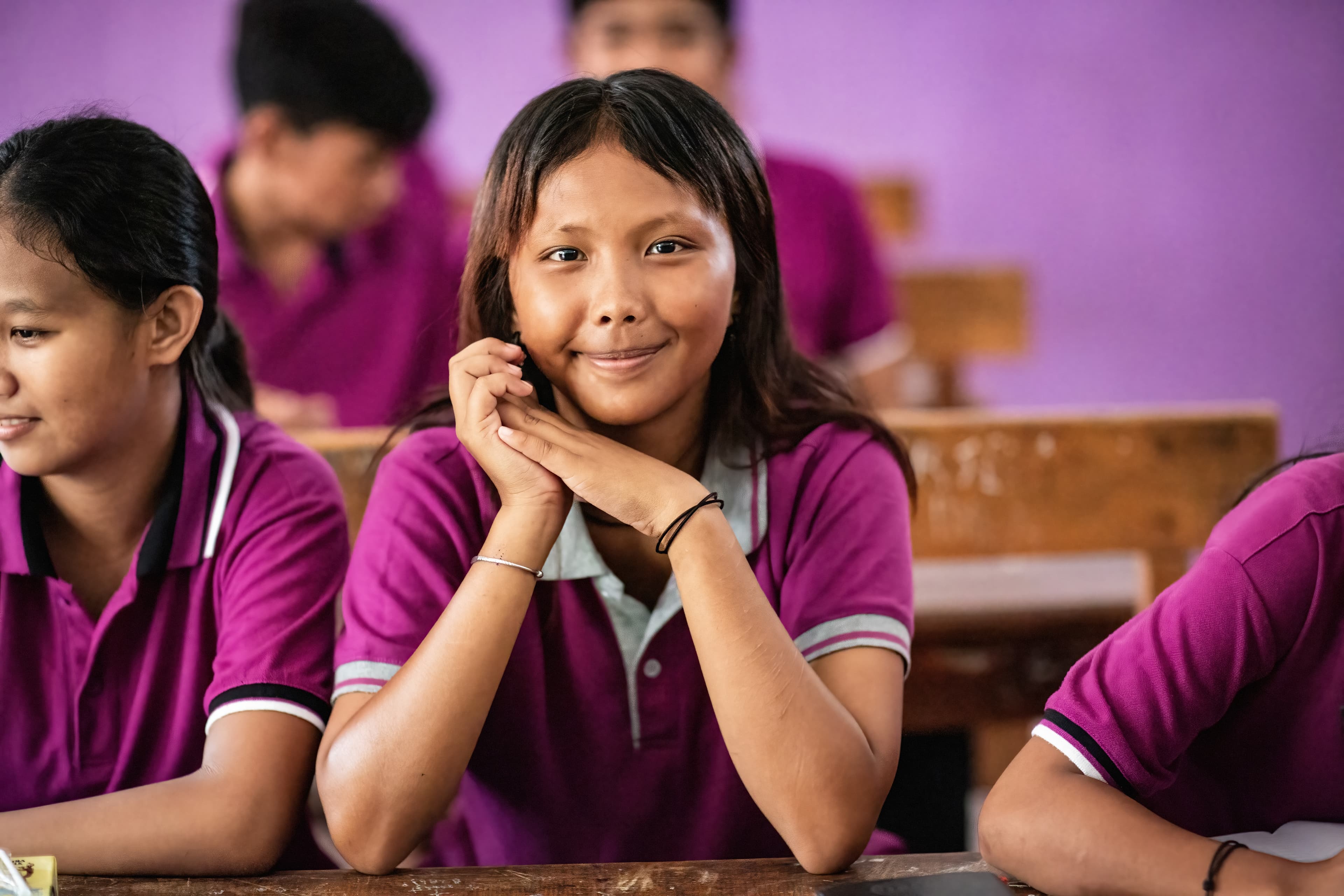 A young girl is sitting at her desk smiling and looking at the camera.