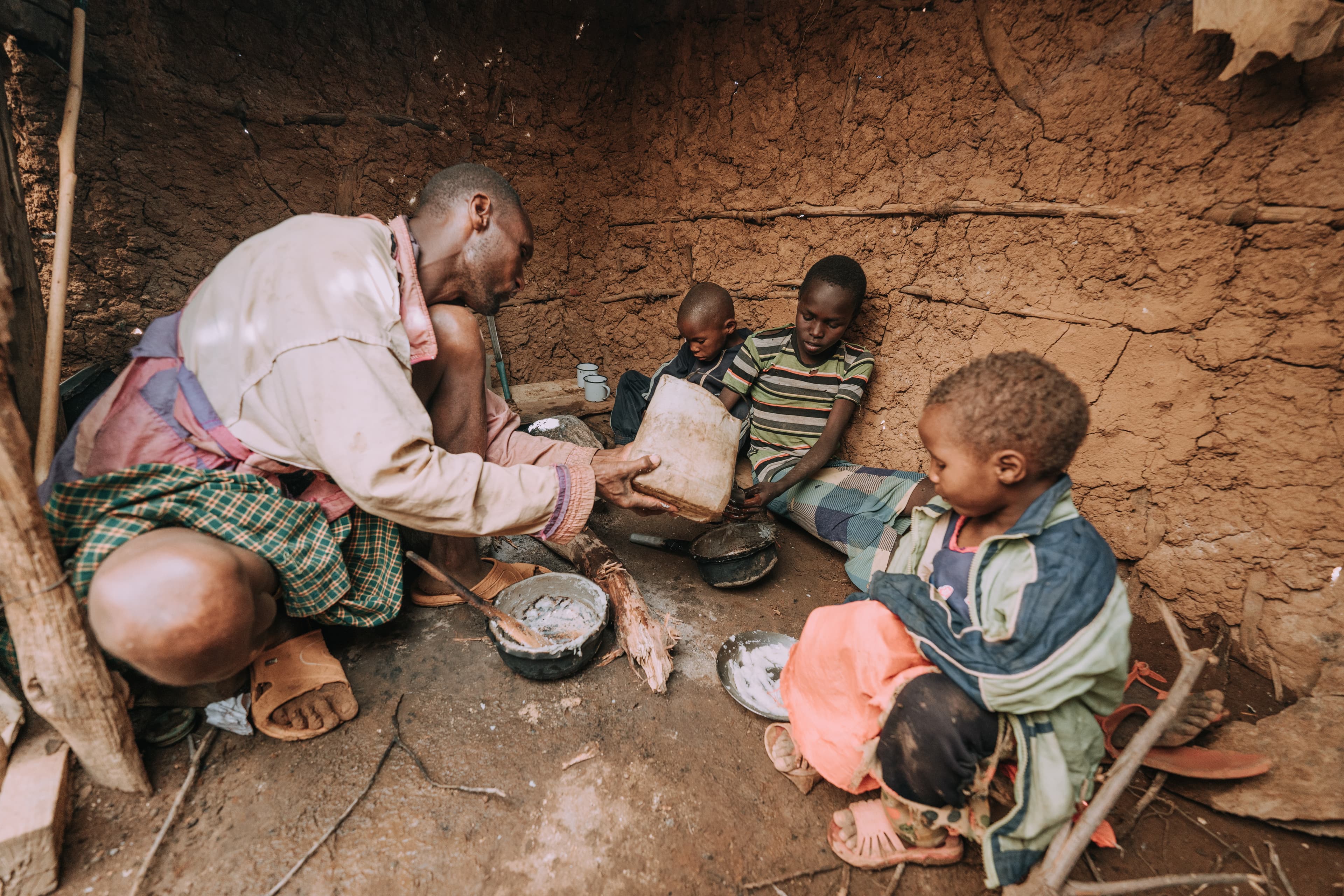 A father is preparing food for his family as he sits on the dirt floor of his home alongside three children.
