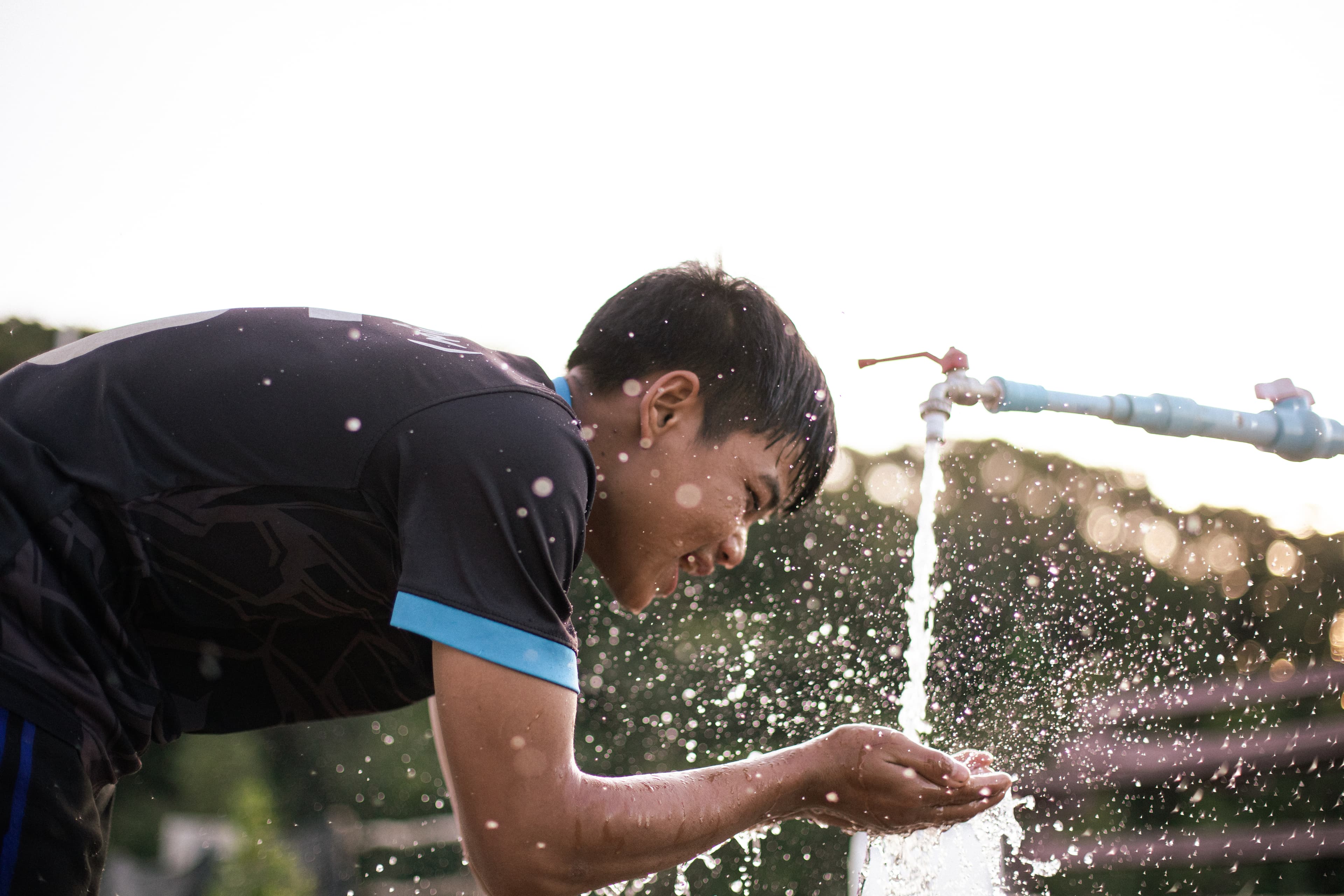 Teenaged boy smiles, splashing water from outdoor faucet on his face.