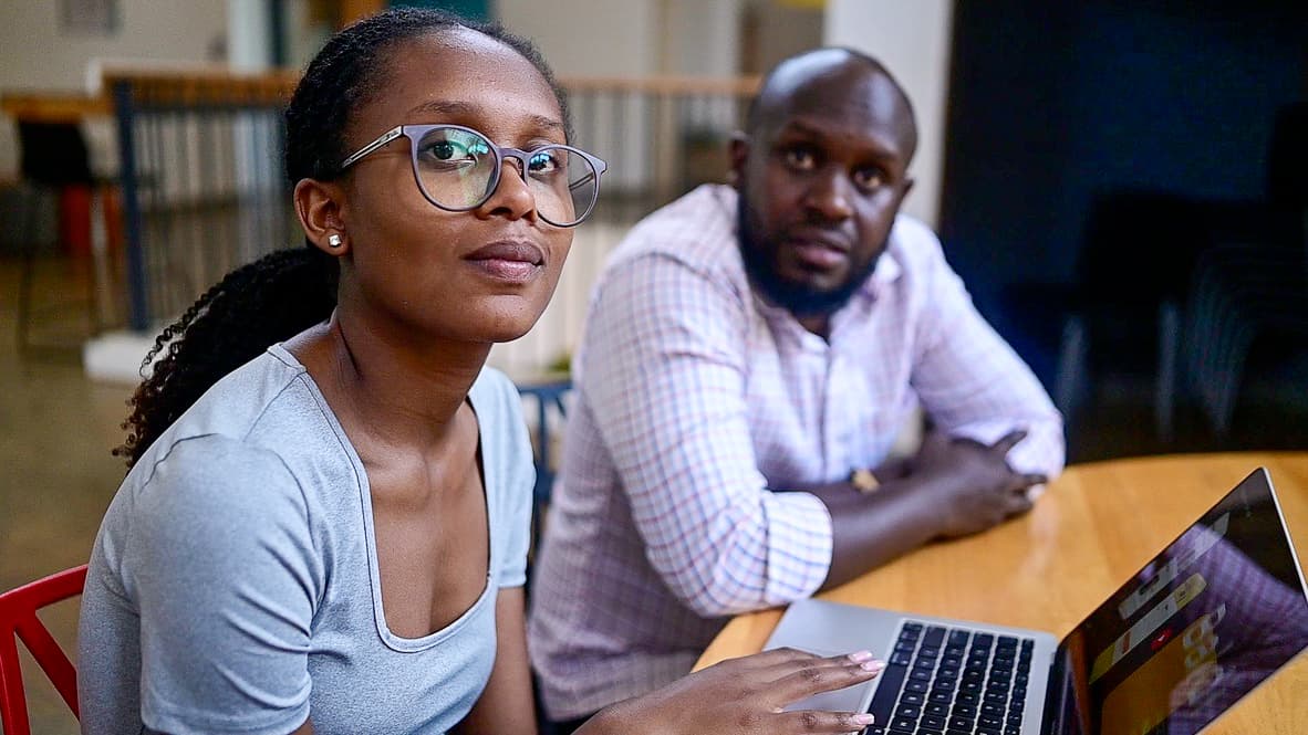 Older teenaged girl looks at the camera with her hand on an open laptop. A man is sitting in the background.