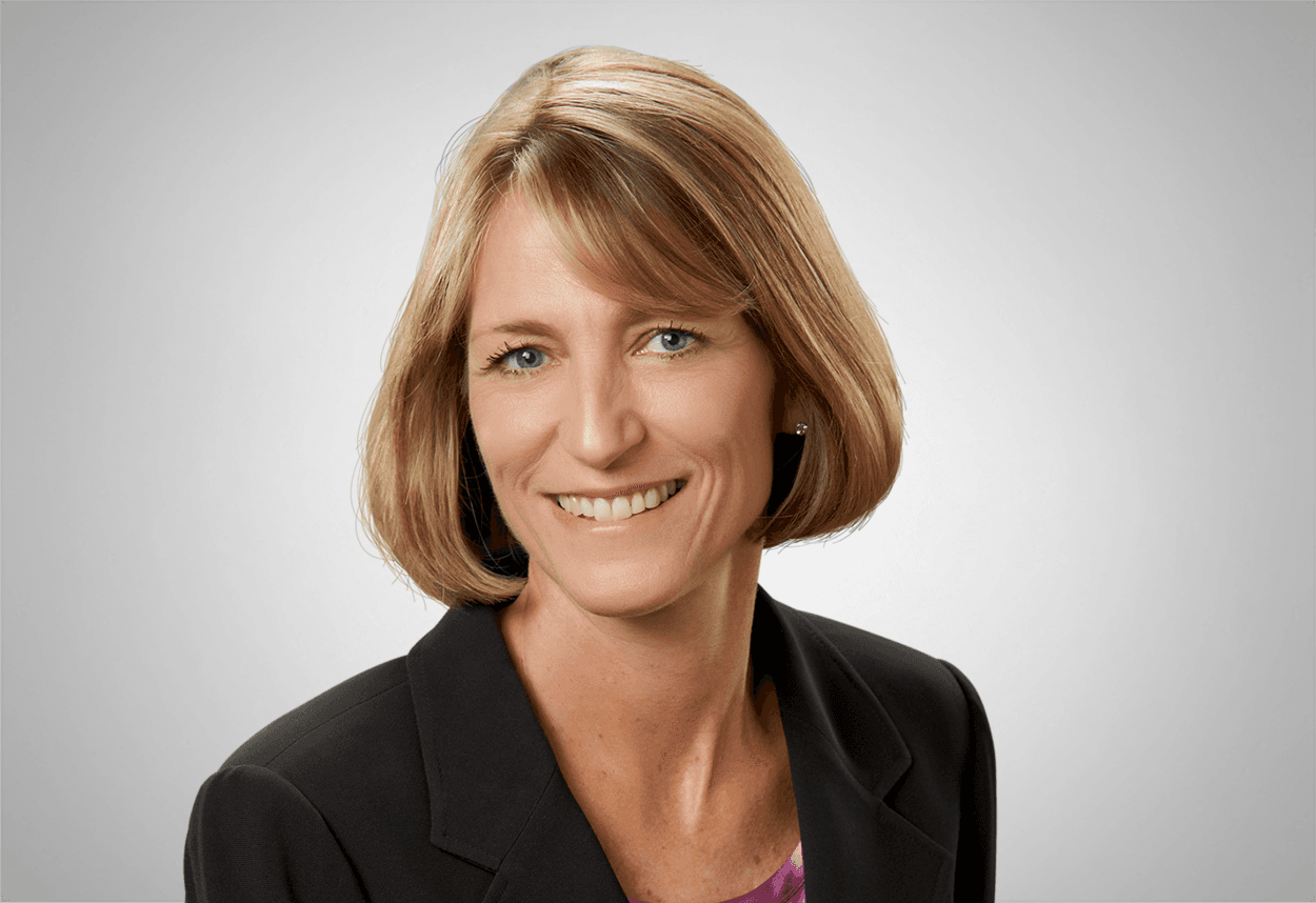 Dr. Judy Briscoe Golz wears a purple top and black blazer is smiling, with a grey background behind her.