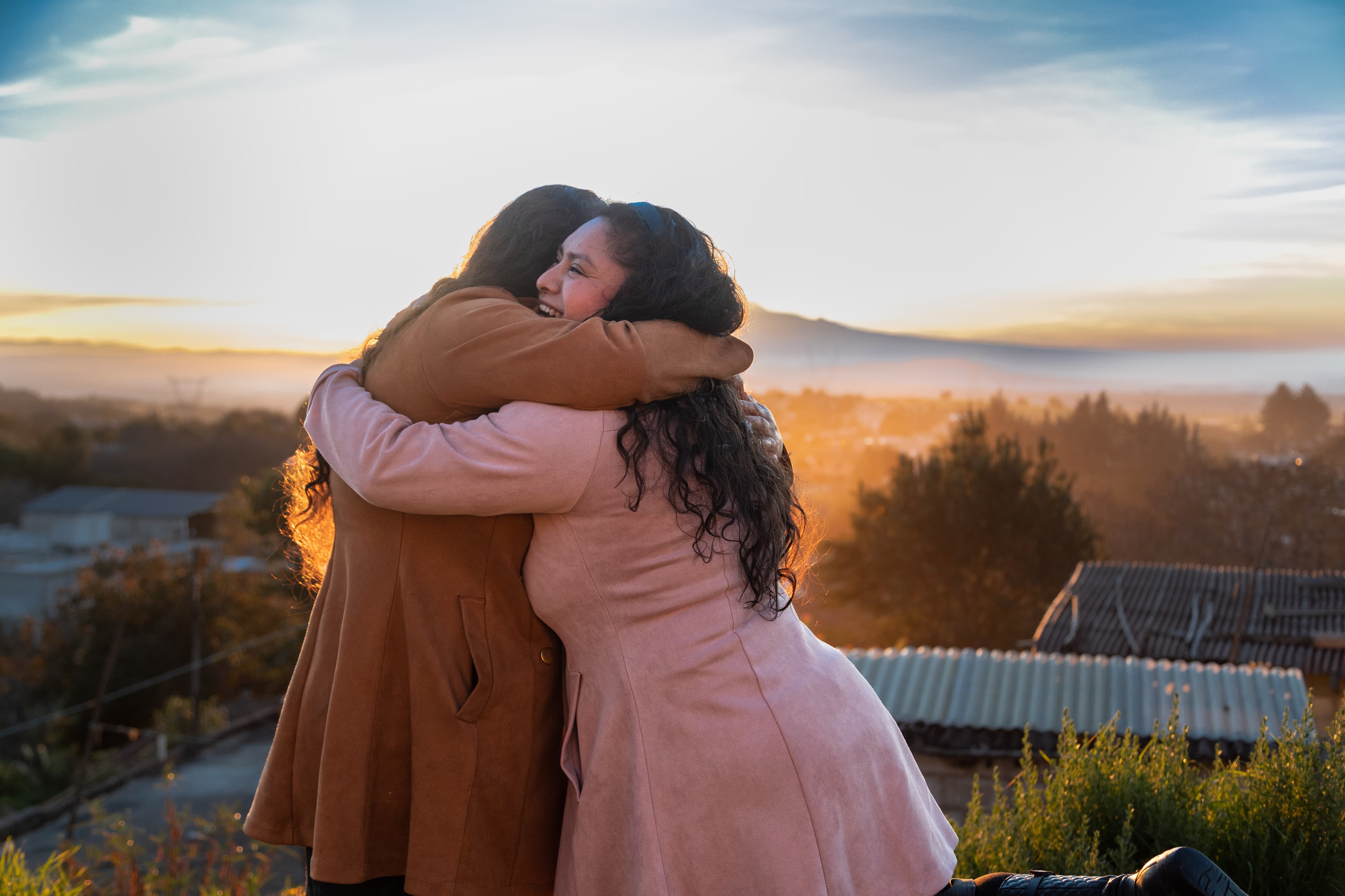 Two women embrace each other in front of a beautiful landscape of mountains, trees and sky.