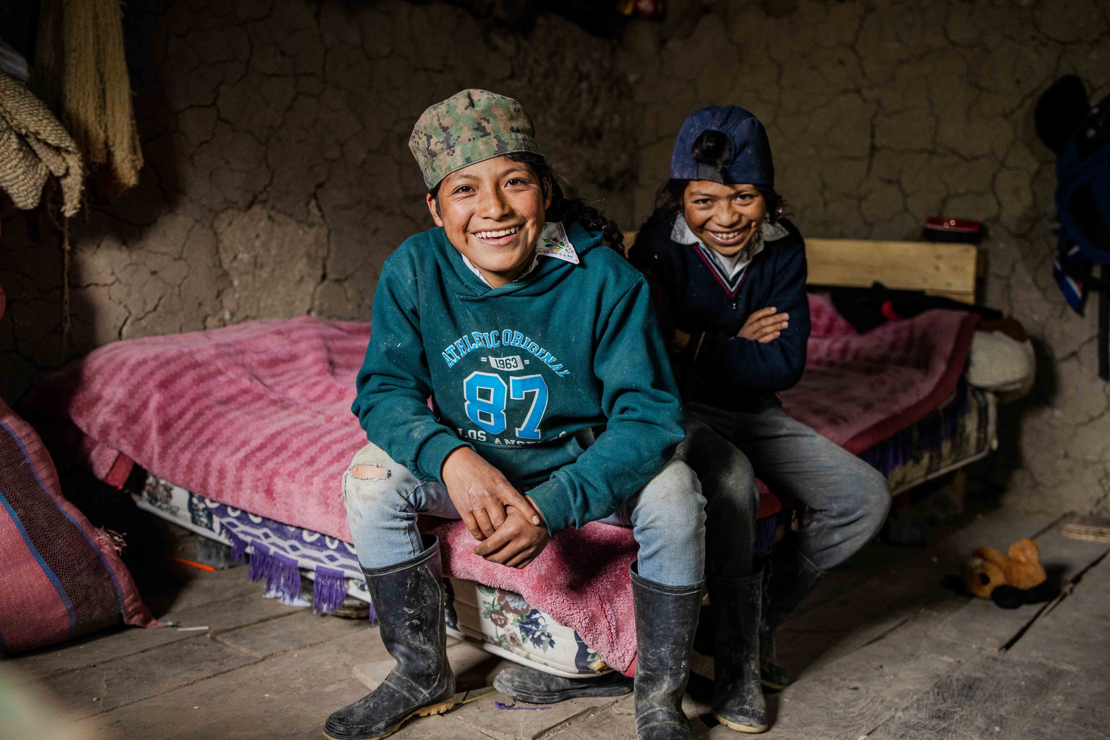 Two Ecuadorean teen boys wearing hats sit on a bed and smile.