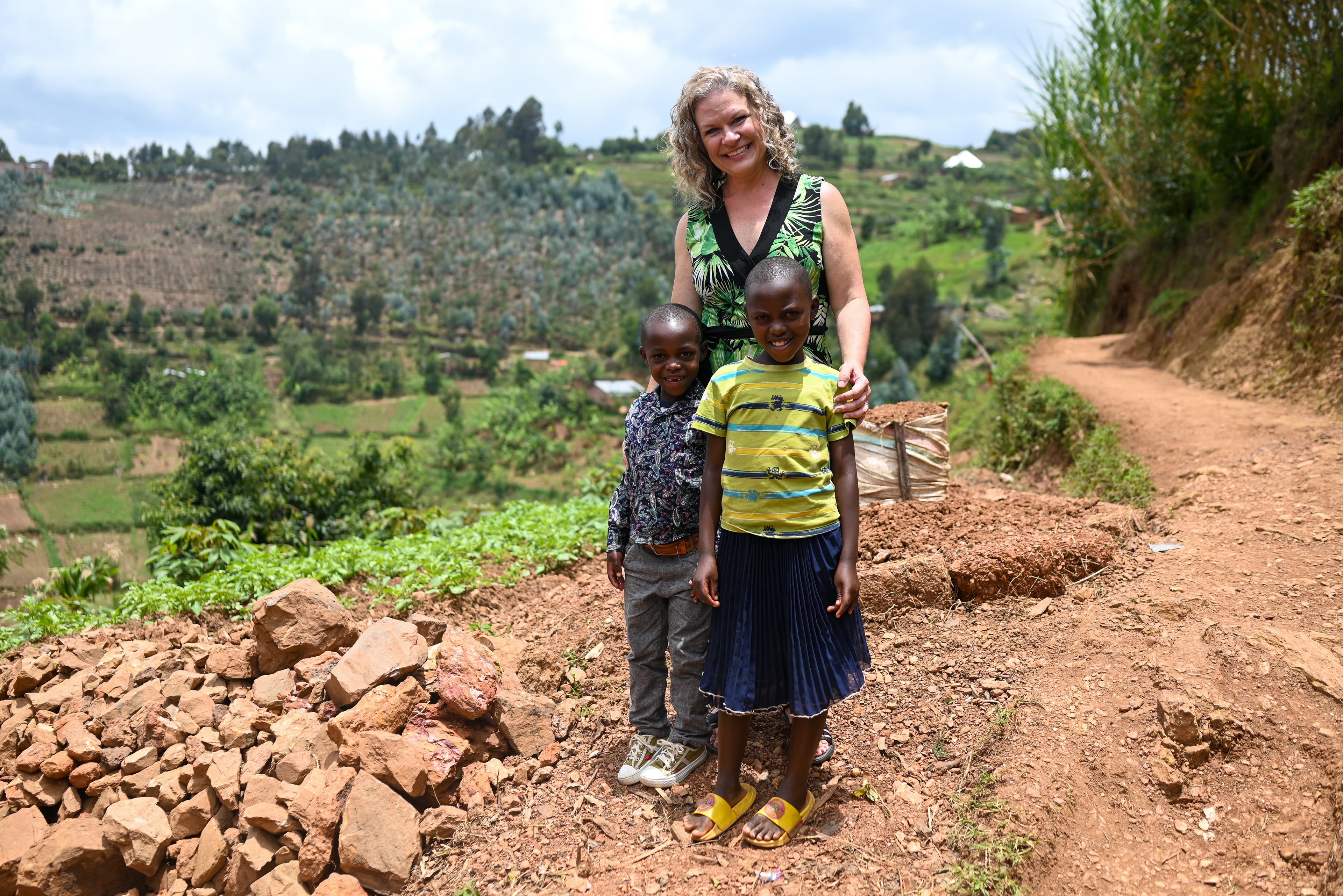 A woman in a green dress stands on a red dirt path with two young children. One child is wearing a yellow shirt and blue skirt, and the other is wearing a blue shirt and gray pants. They are all smiling, and foliage and farms are behind them.