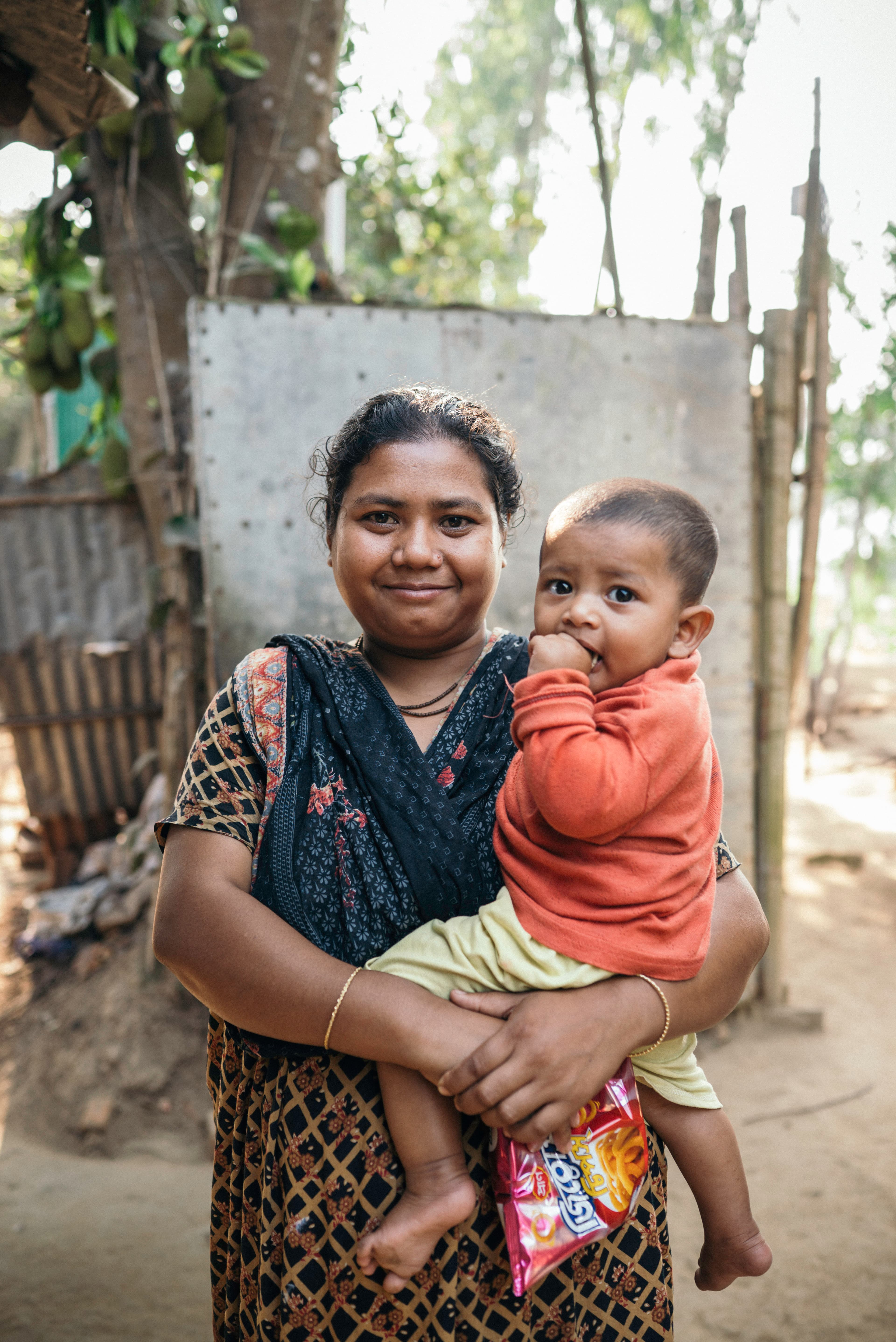 A mother holds her infant son in her arms outside her home, smiling, with trees and vegetation in the background.