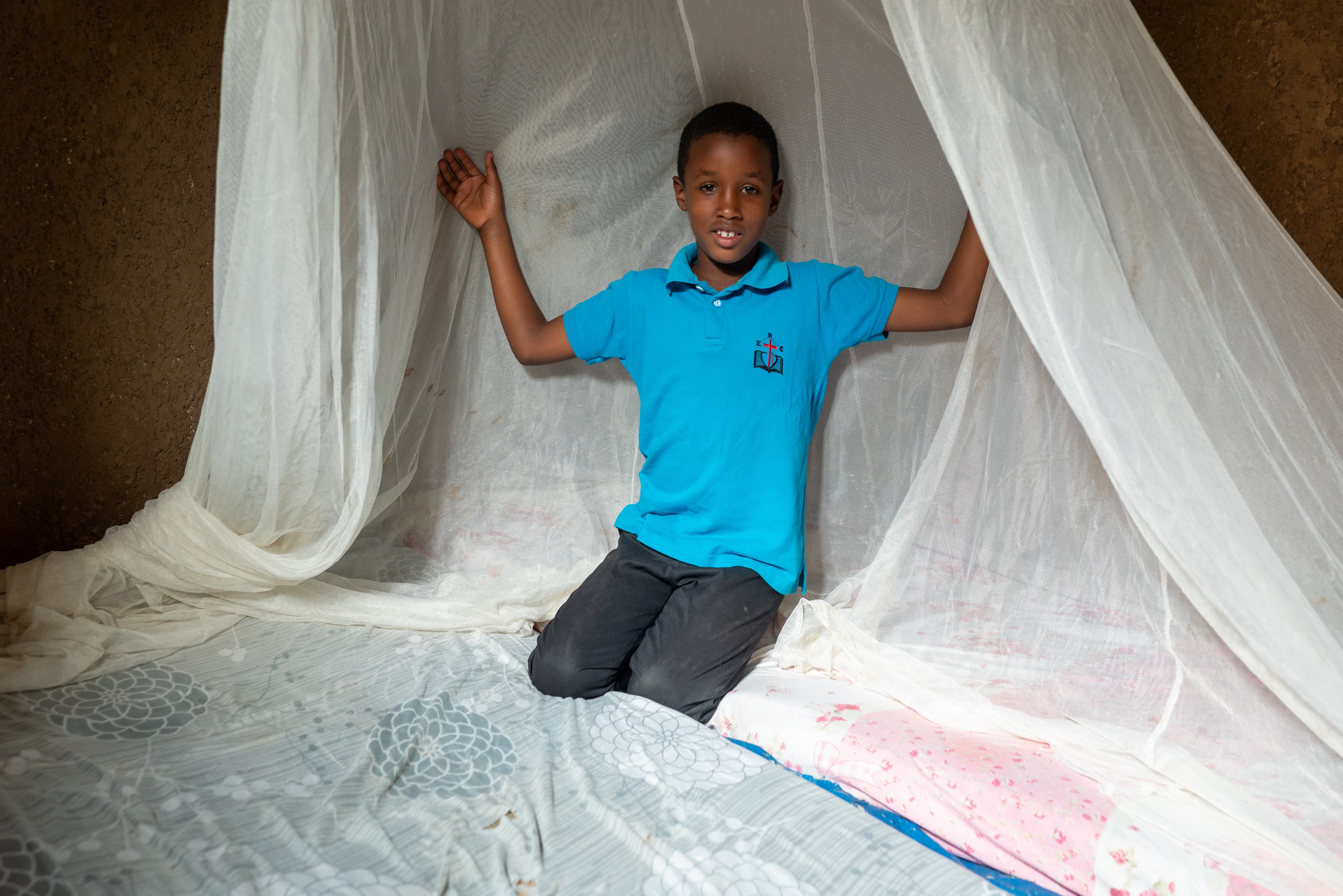 A young boy wearing a blue polo kneels on his bed while smiling under a mosquito net.