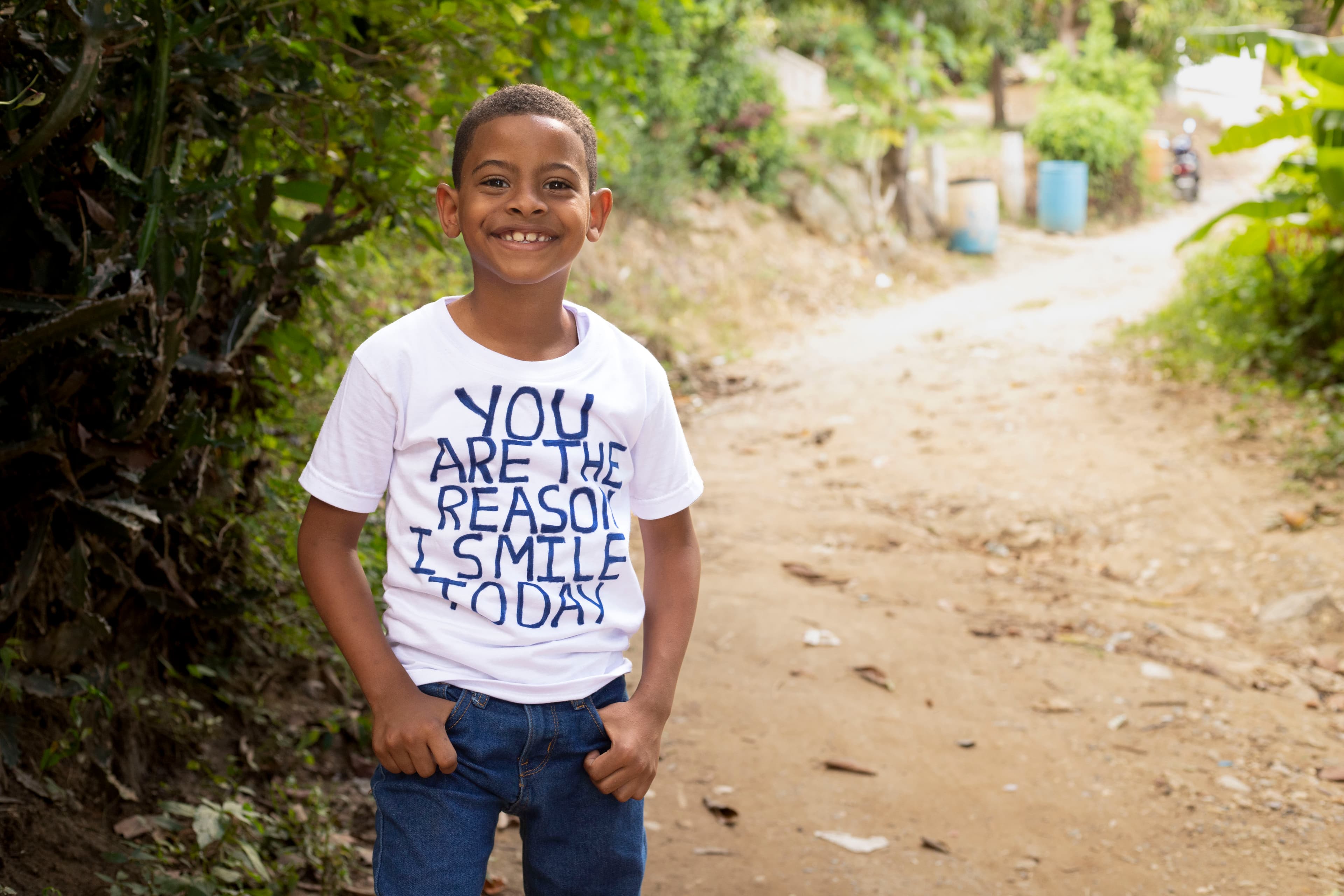 A young boy smiles for the camera wearing a shirt that says, “You are the reason I smile today.”