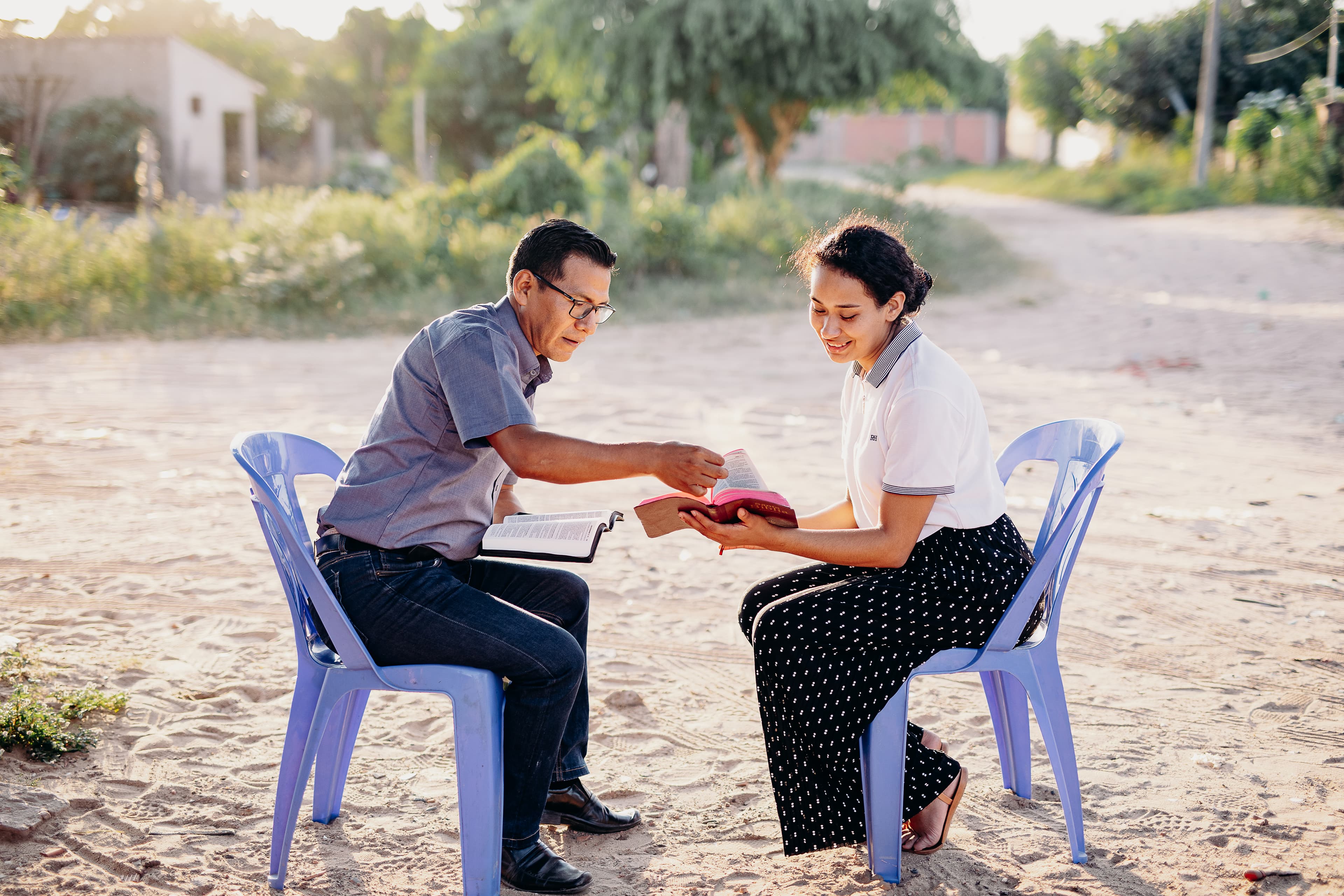 A teen Bolivian girl and an older man sit on blue chairs while reading the Bible.