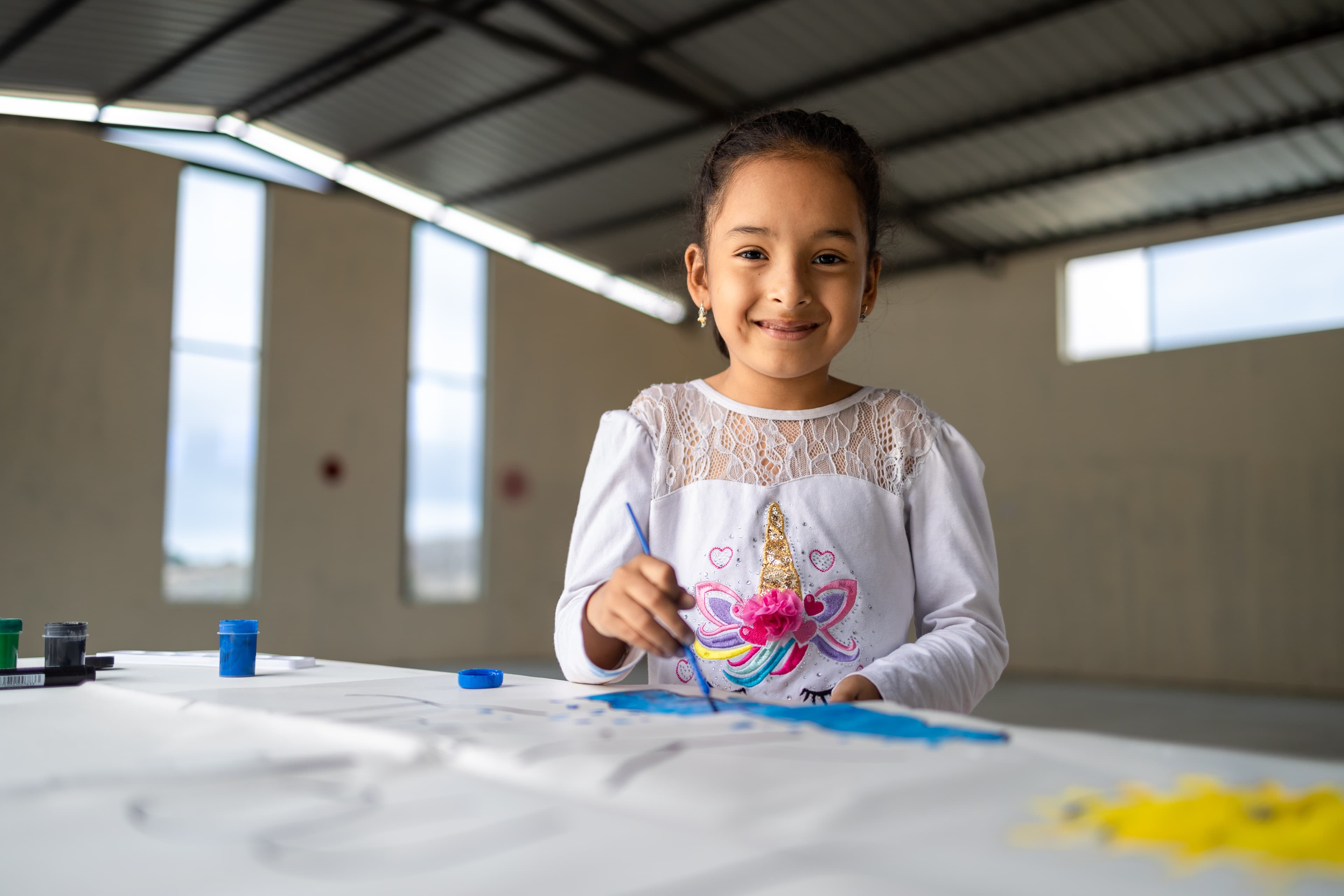 Young girl from Ecuador smiles at the camera while painting at a table.