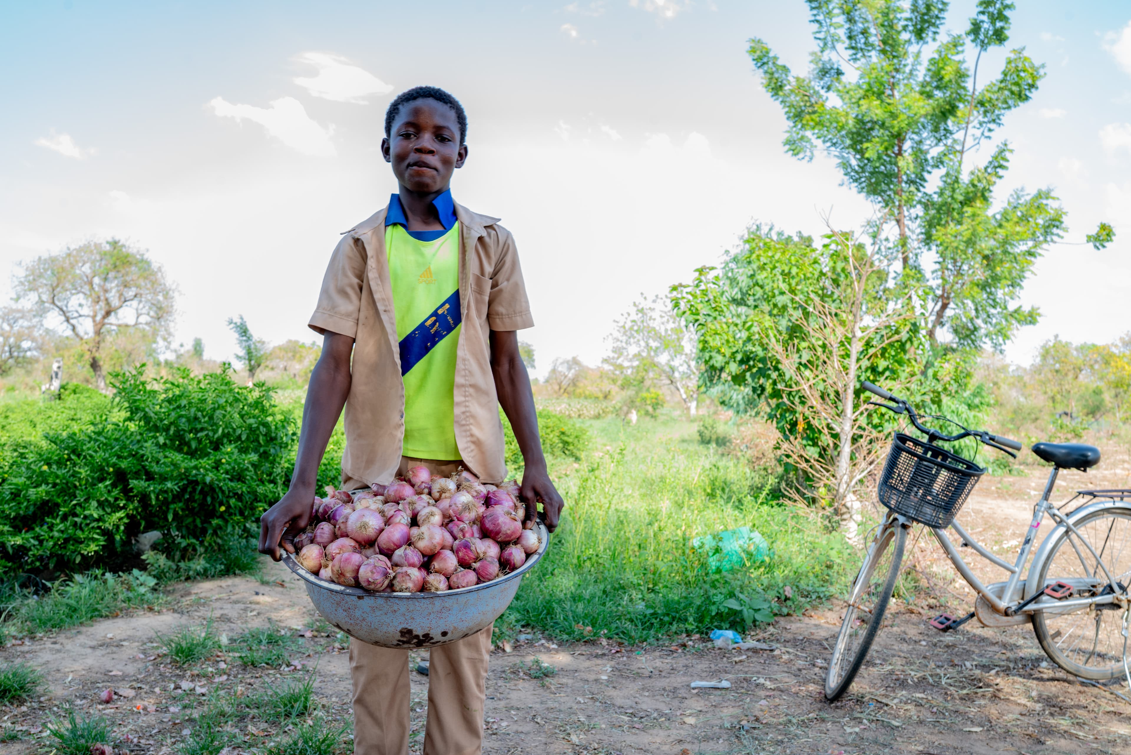A teen African boy smiles for the camera while holding a metal tub of fresh onions.