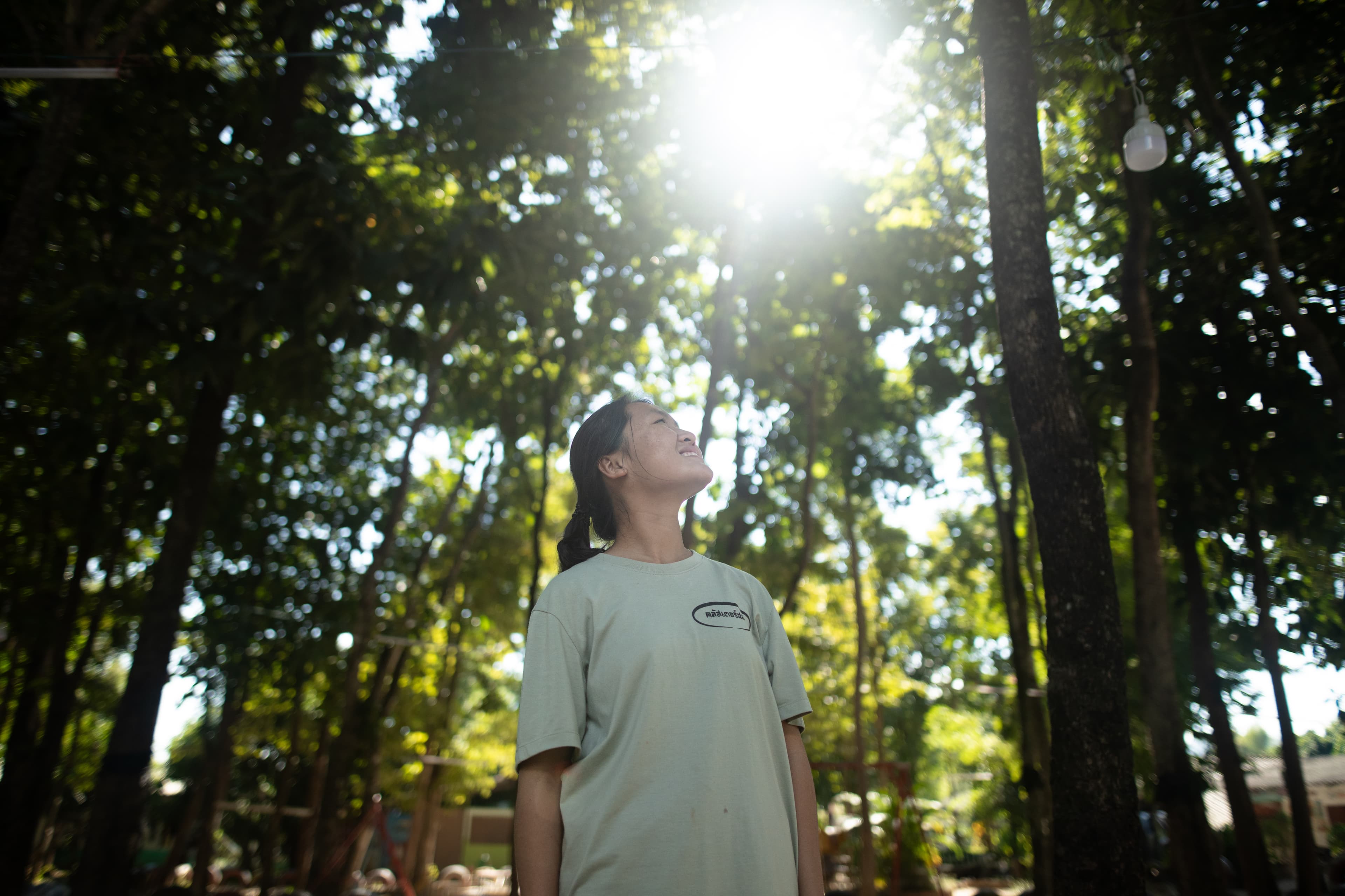 A teen Thai girl looks up into the trees with the sun shining on her face.