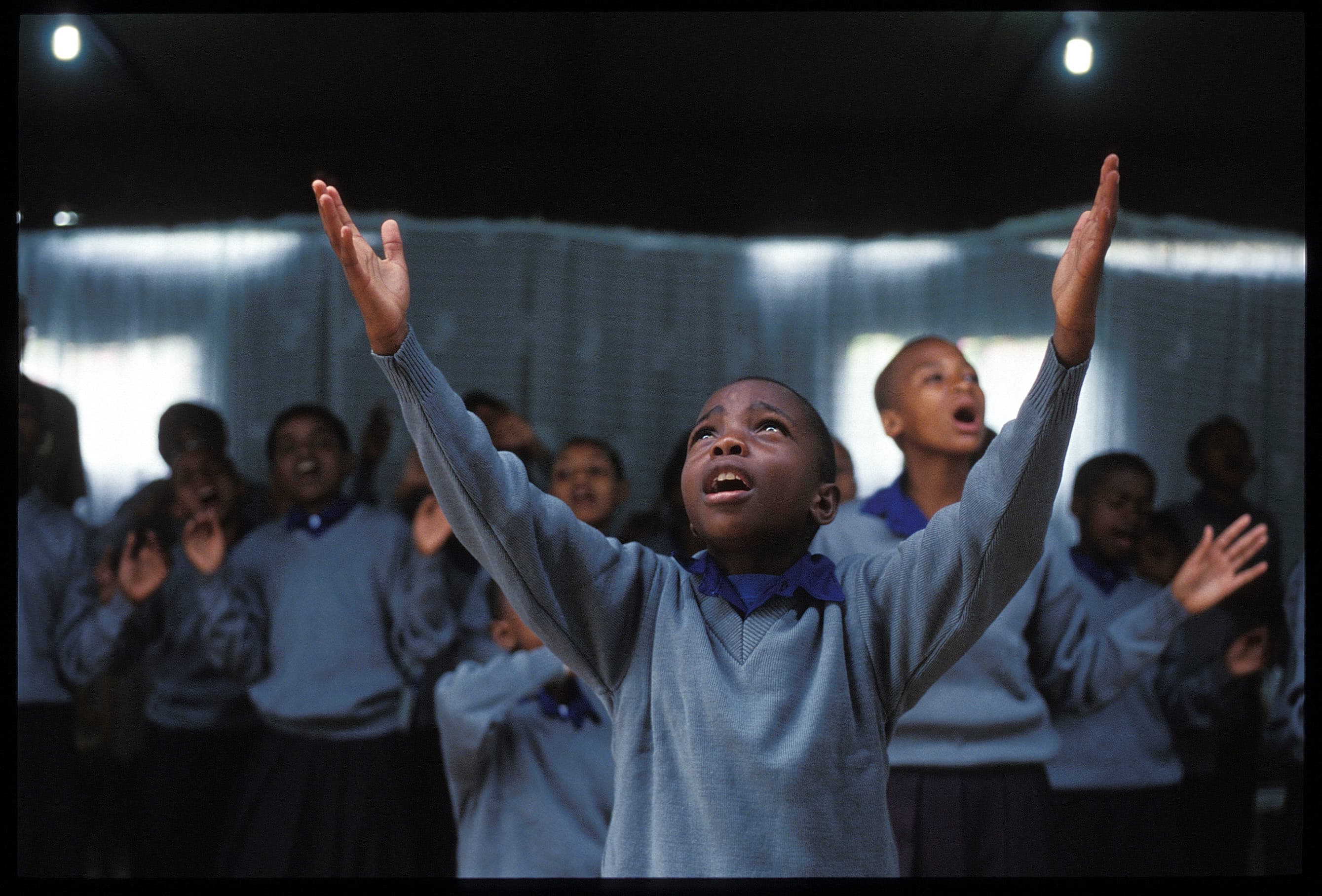 A young boy raises his hands in worship while singing, looking upward. Other children sing together in the background.