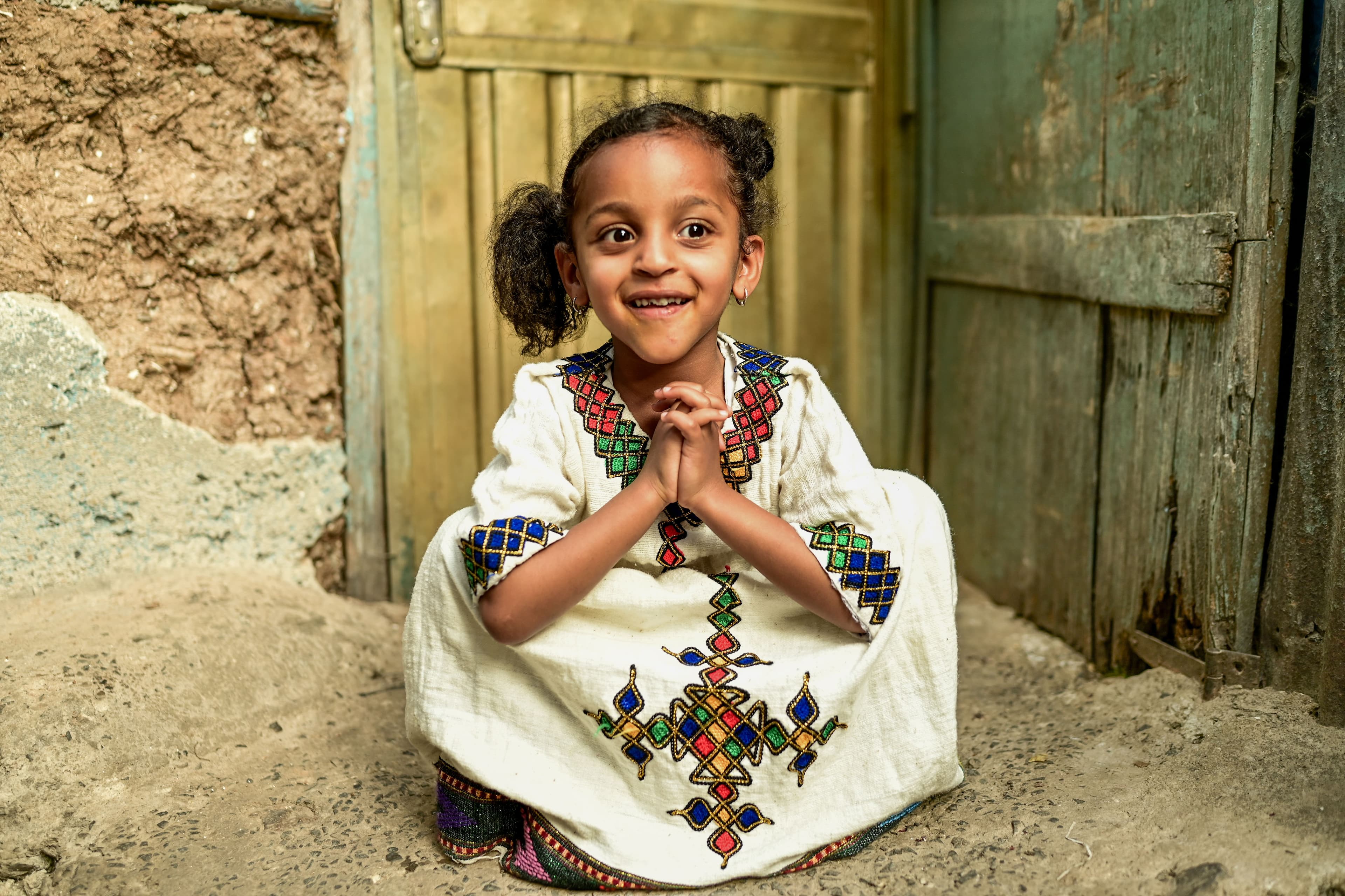 A young girl sits on the ground folding her hands together and smiling.