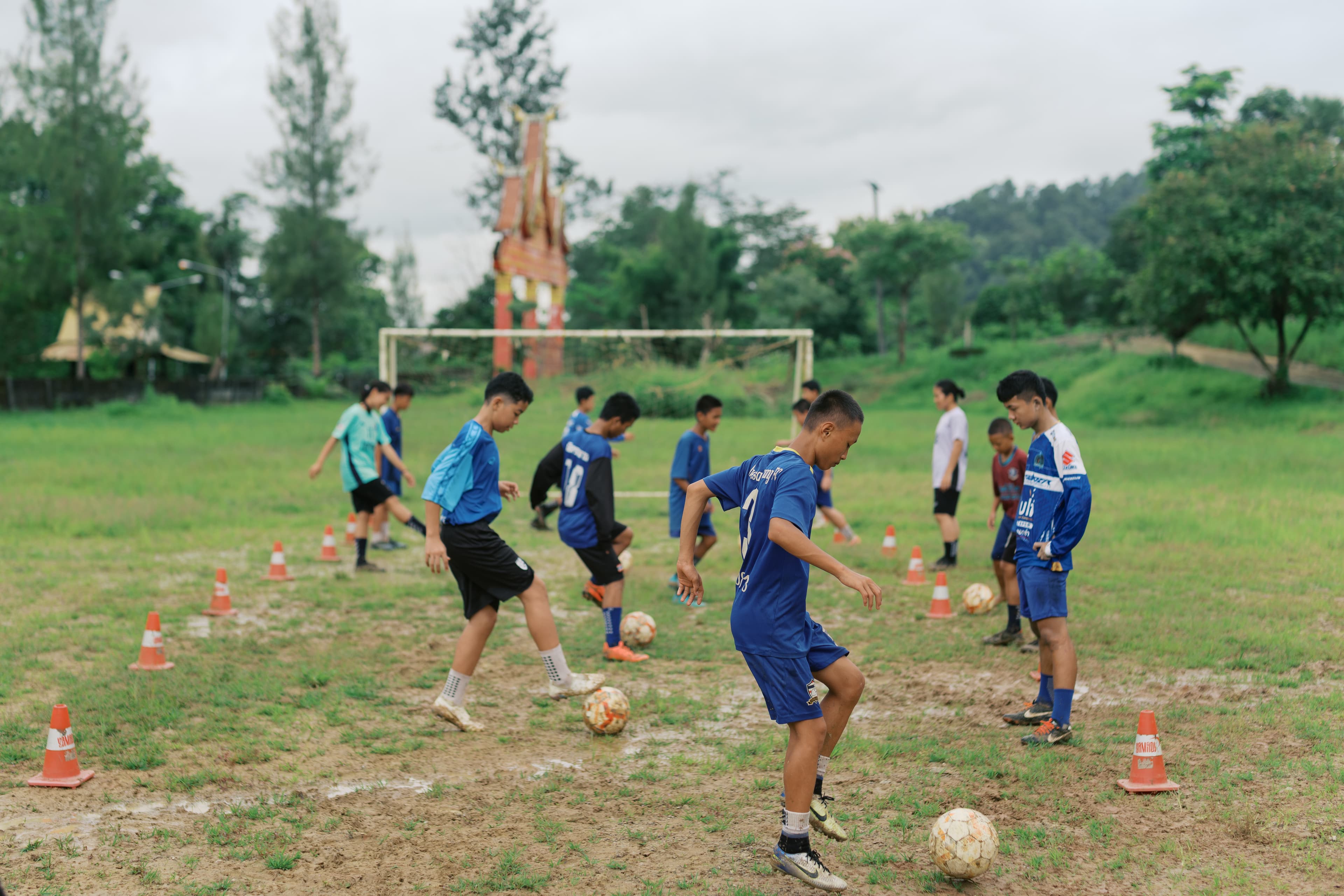 A group of youth play football on a grassy field with trees and buildings in the background.