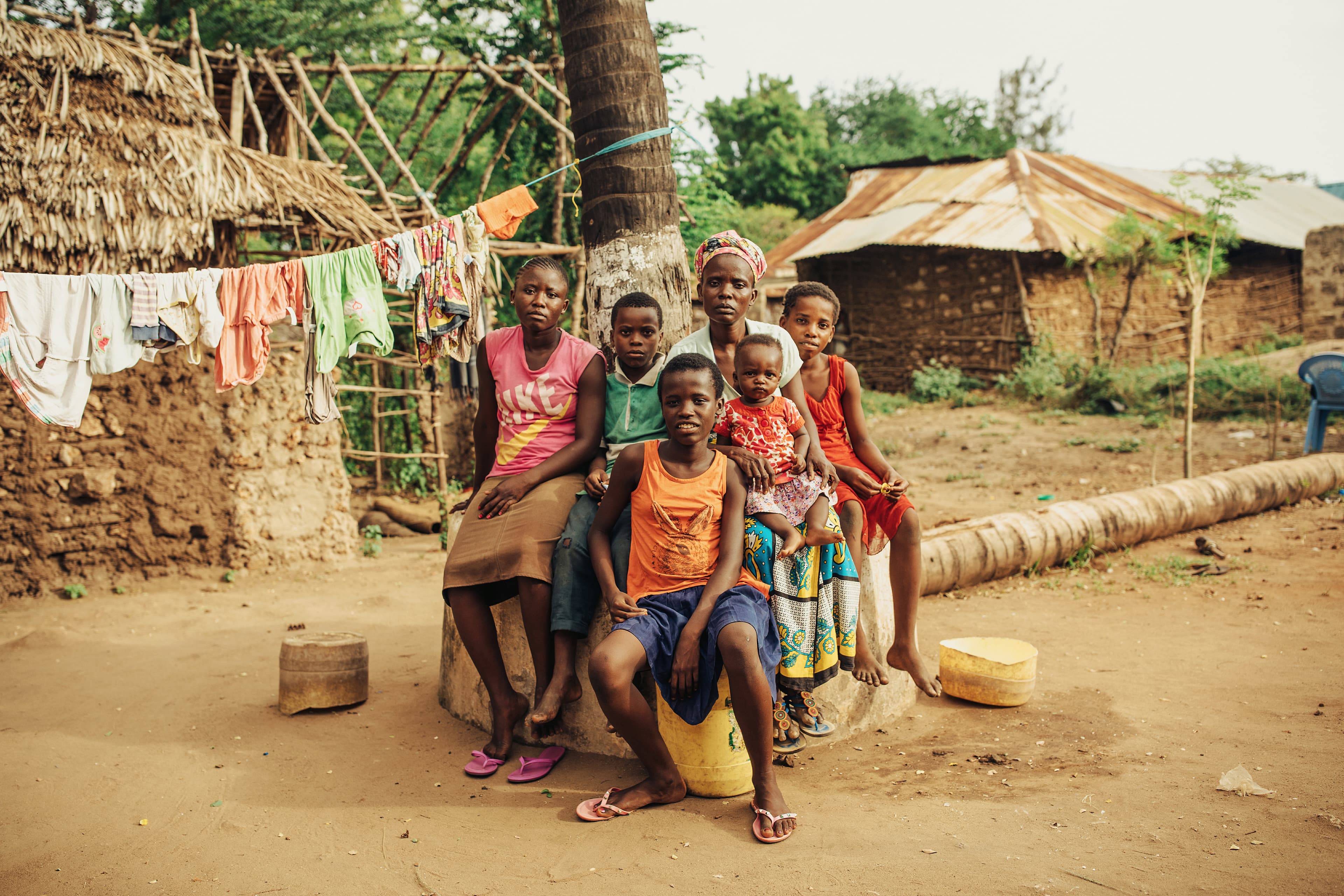 Family of six sits near a tree outside their home in Kenya. The terrain around them is dry.