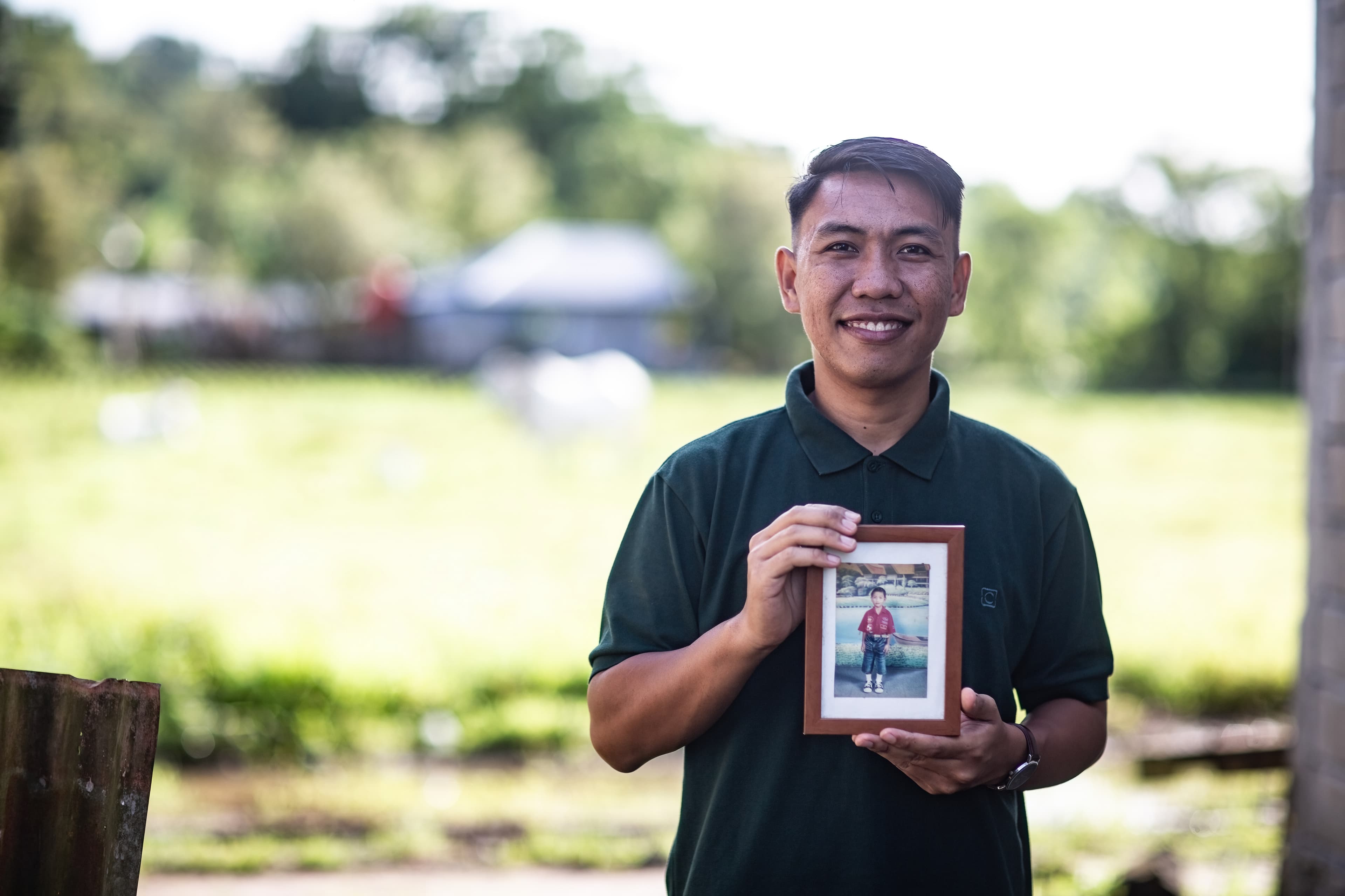 A young man wearing a green polo smiles and holds a photo of himself as a child.