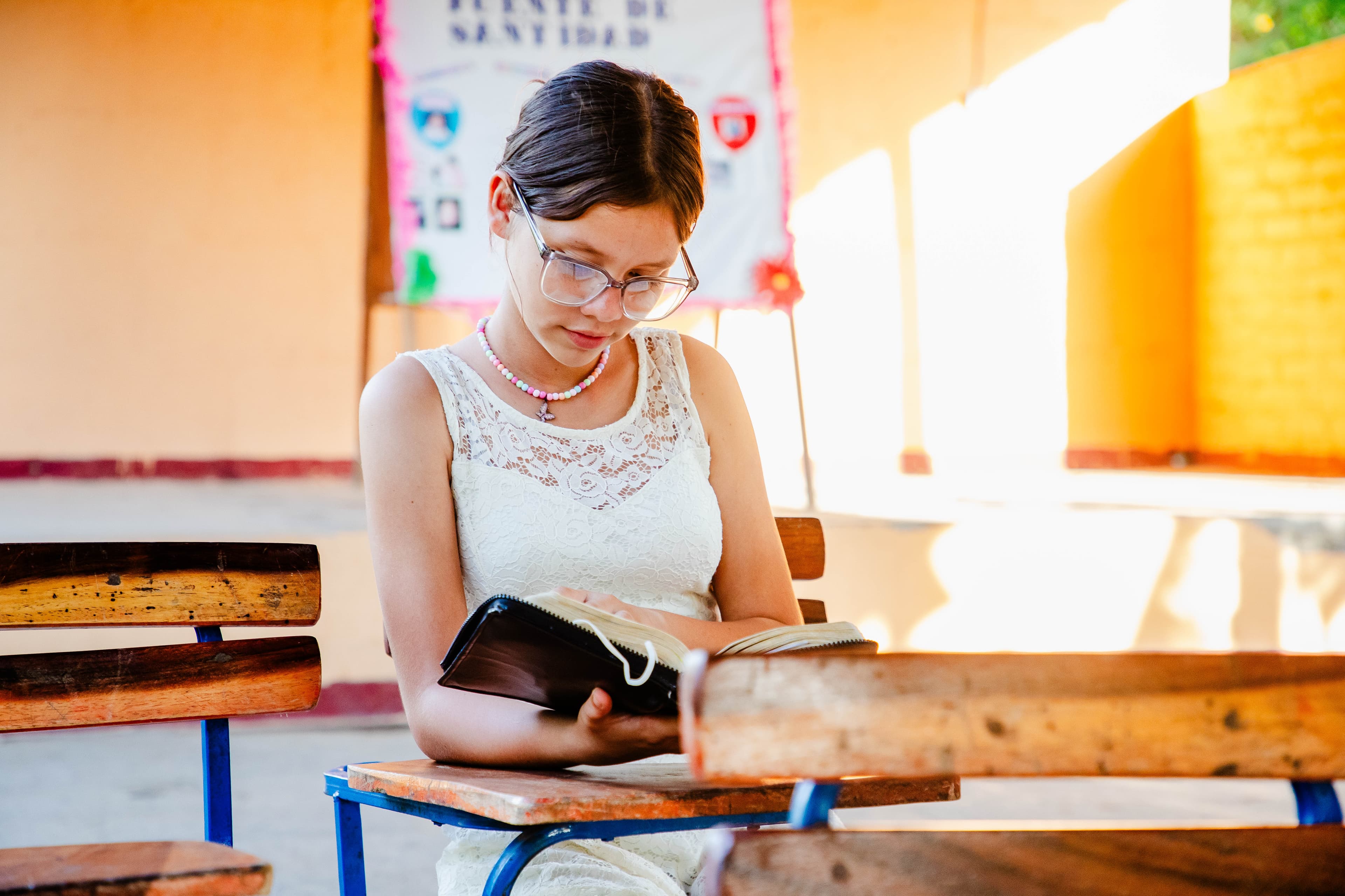 A young girl wearing glasses sits at a desk while reading the Bible.