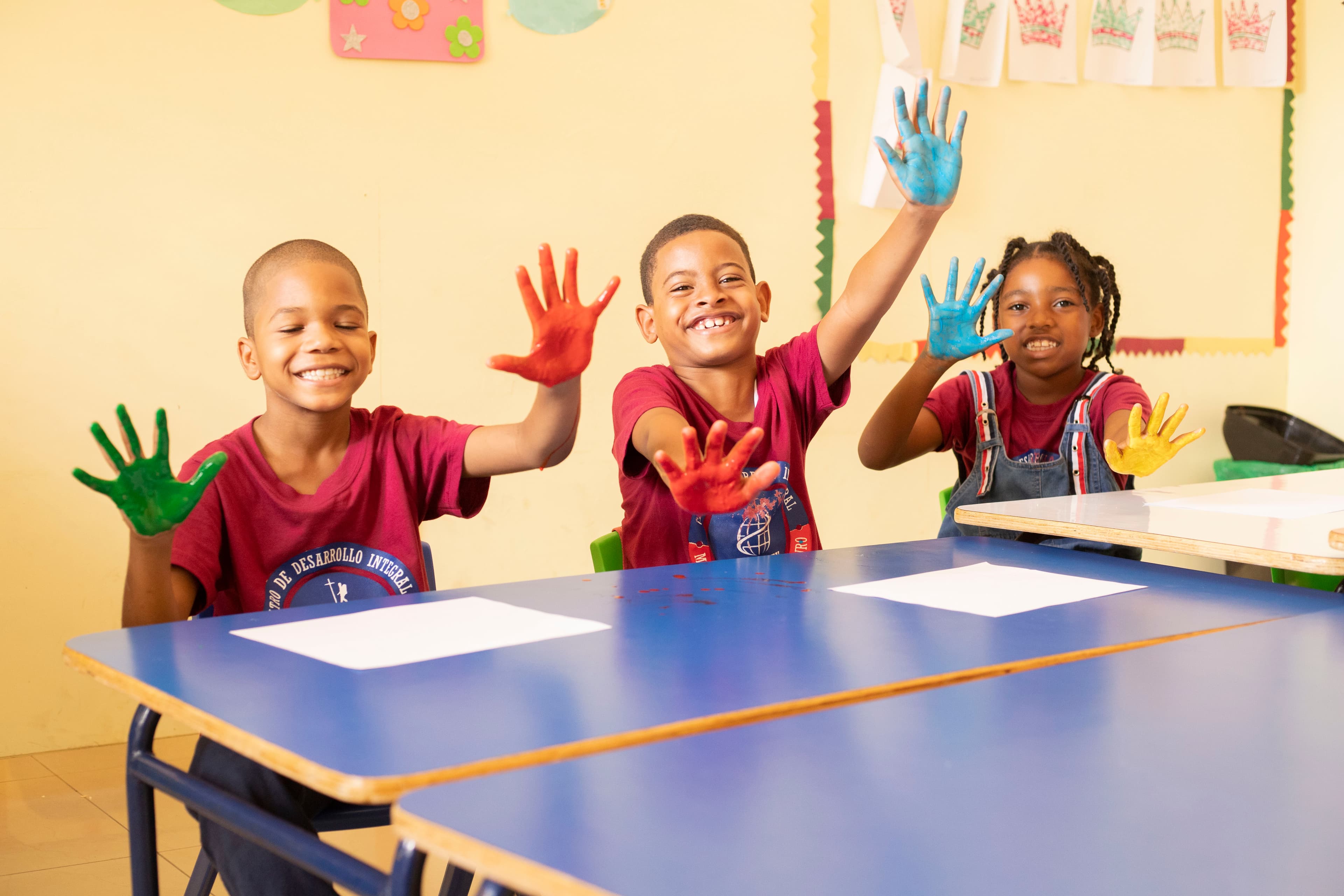 Three young children are sitting at a desk smiling and raising their hands in the air during a craft activity.
