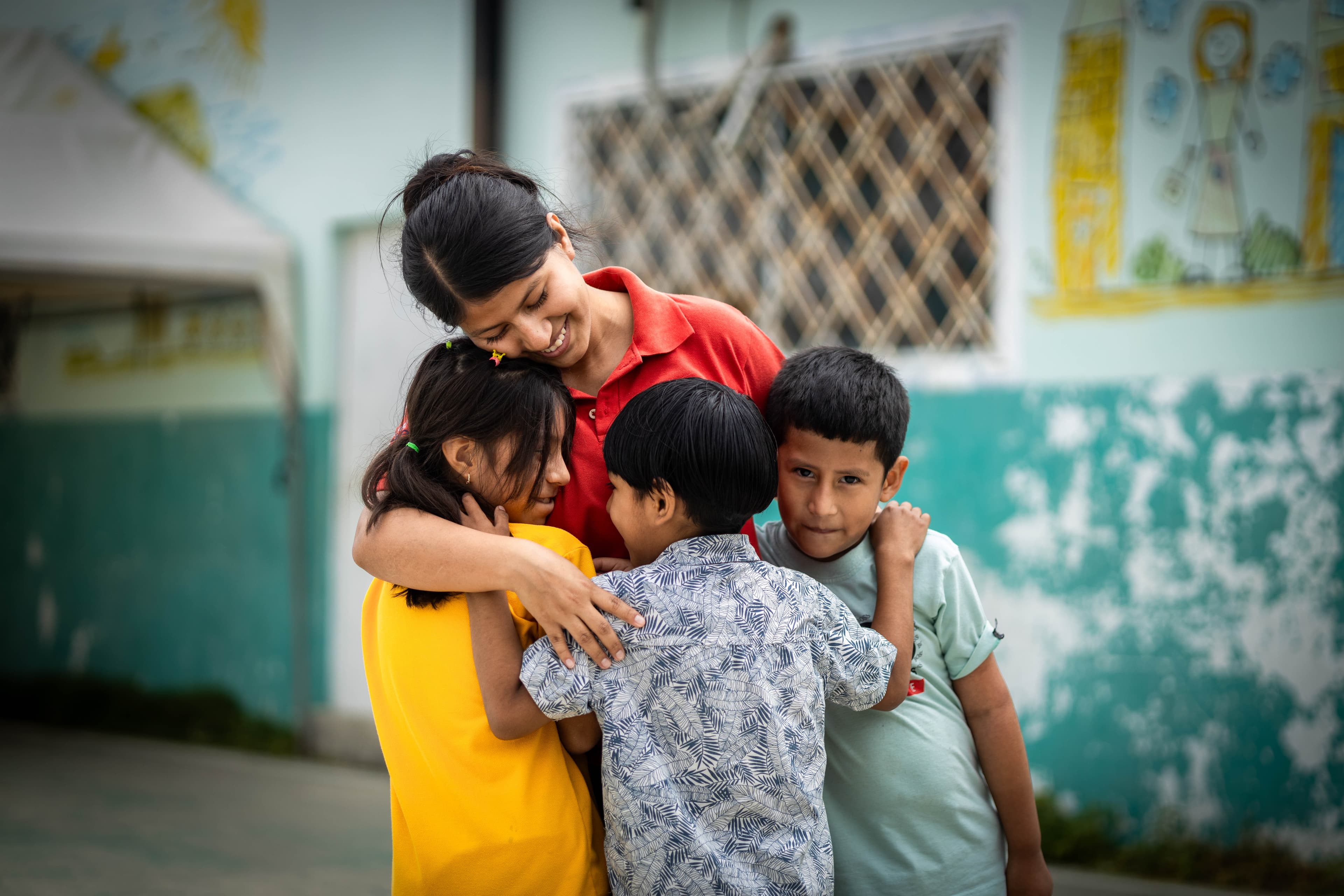 A young woman in a red shirt embraces three children, standing outside in a courtyard, with a blue wall and painted mural in the background.