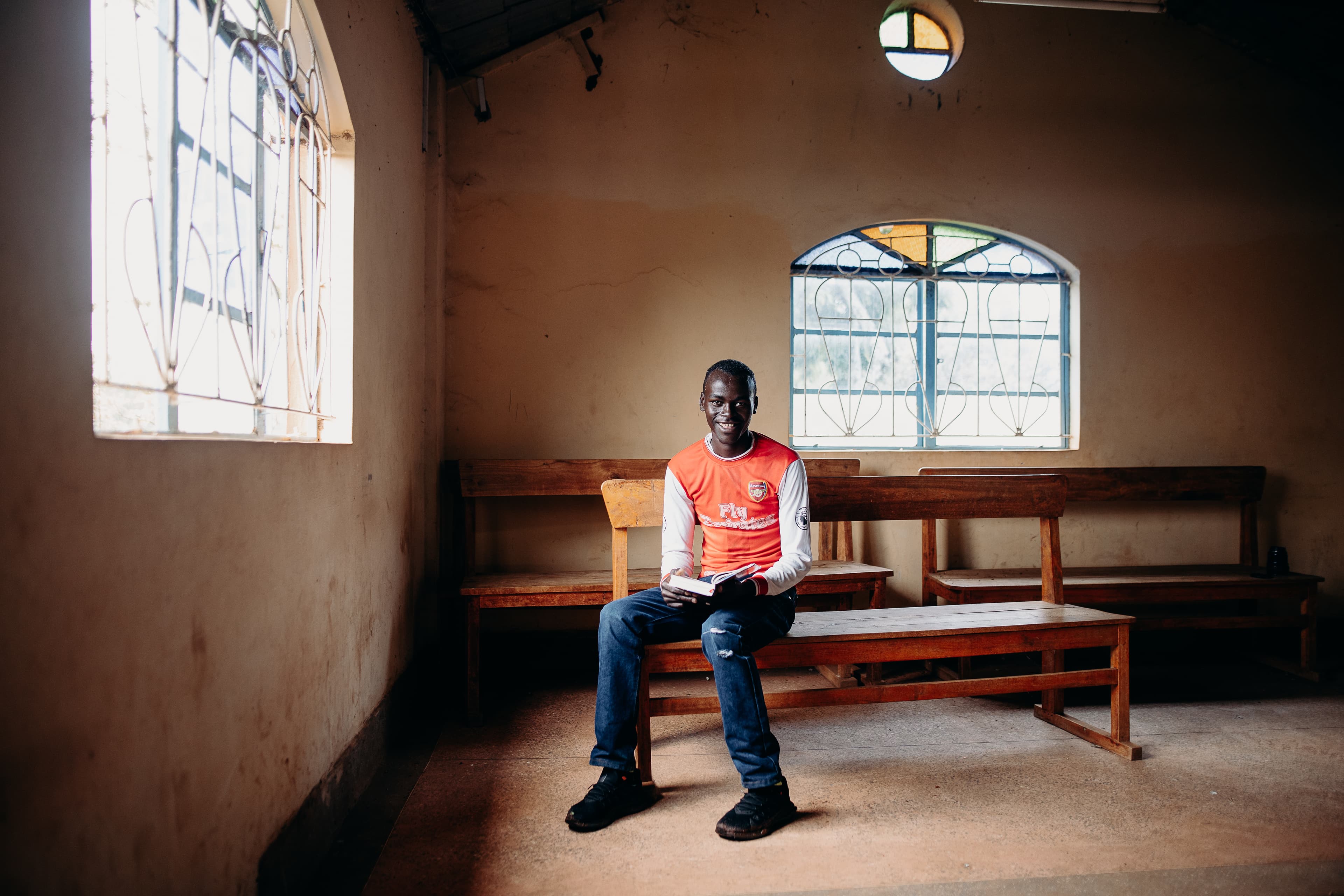 A young African man sits on a wooden bench with a Bible and smiles for the camera.