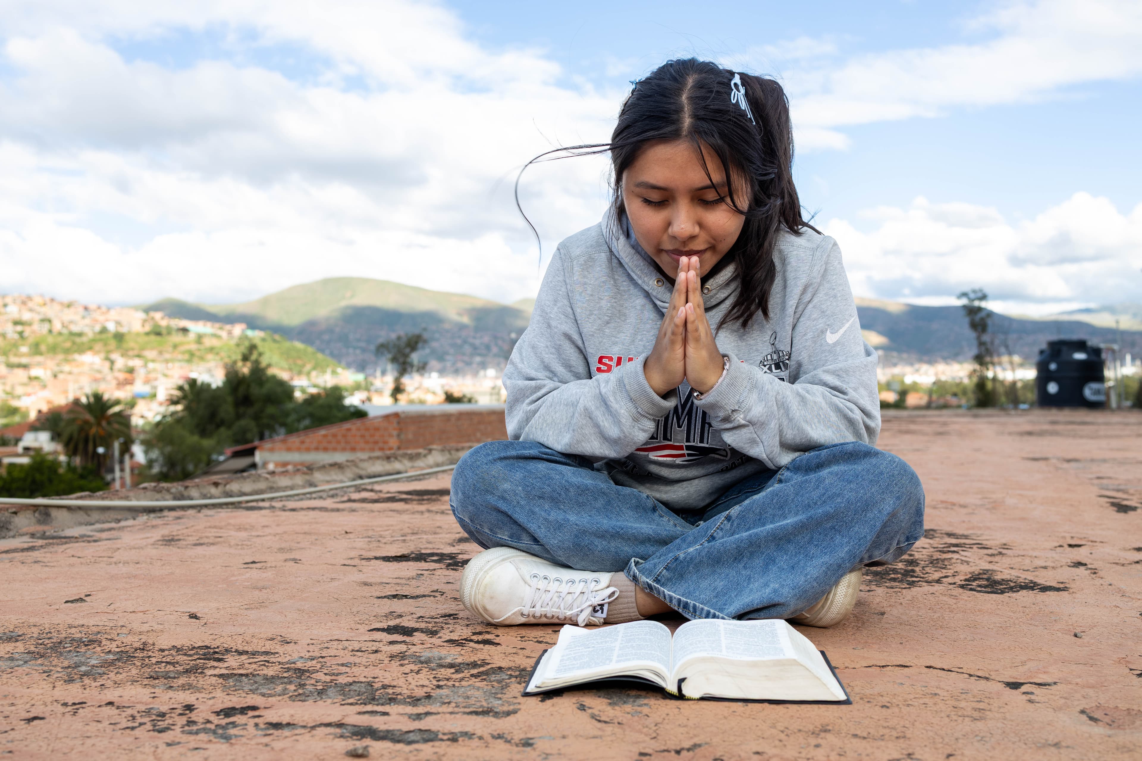 A young girl wearing a gray hoodie prays while sitting in front of her Bible.