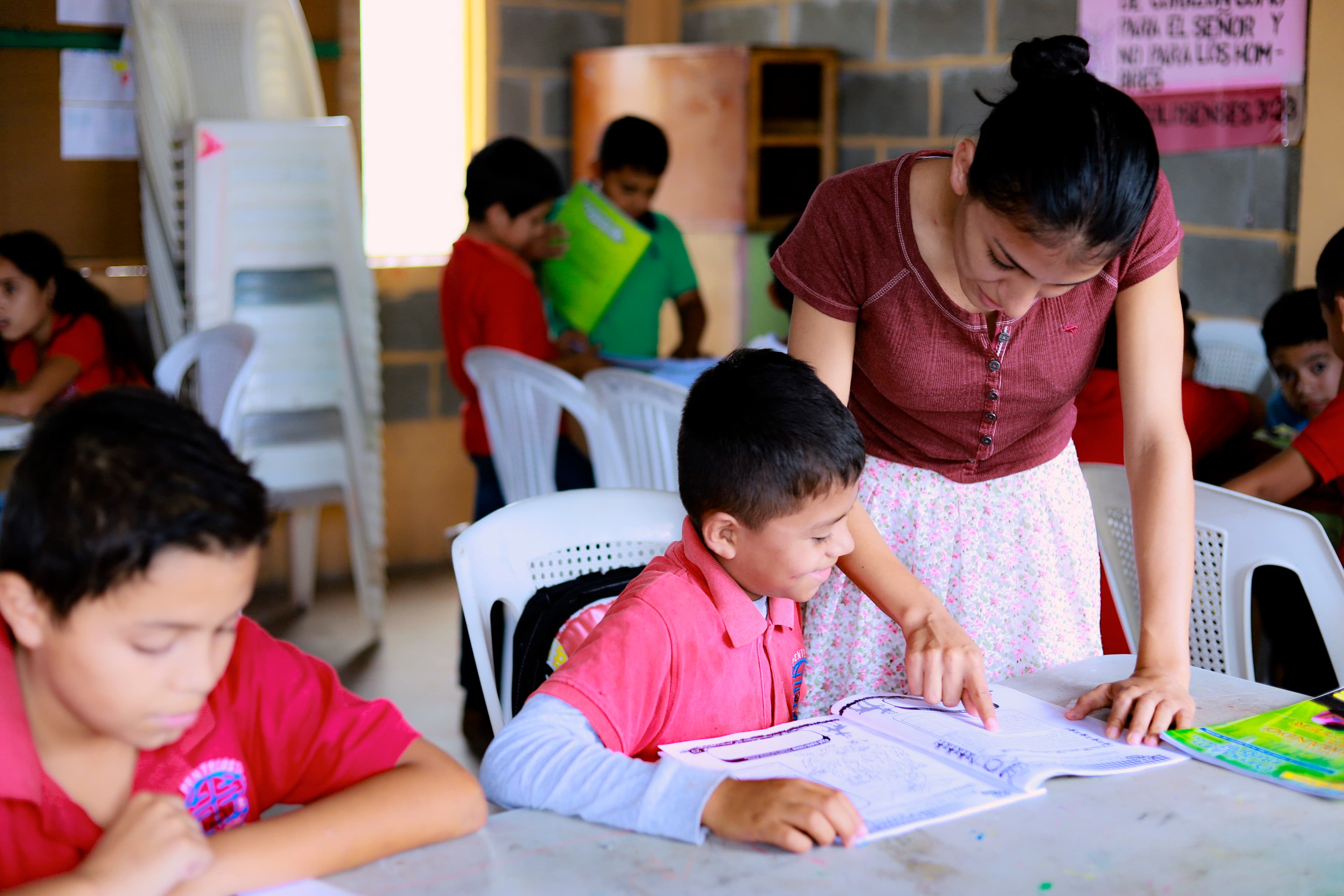 A young woman wearing a red top and pink floral skirt assists a boy reading a workbook at a table.