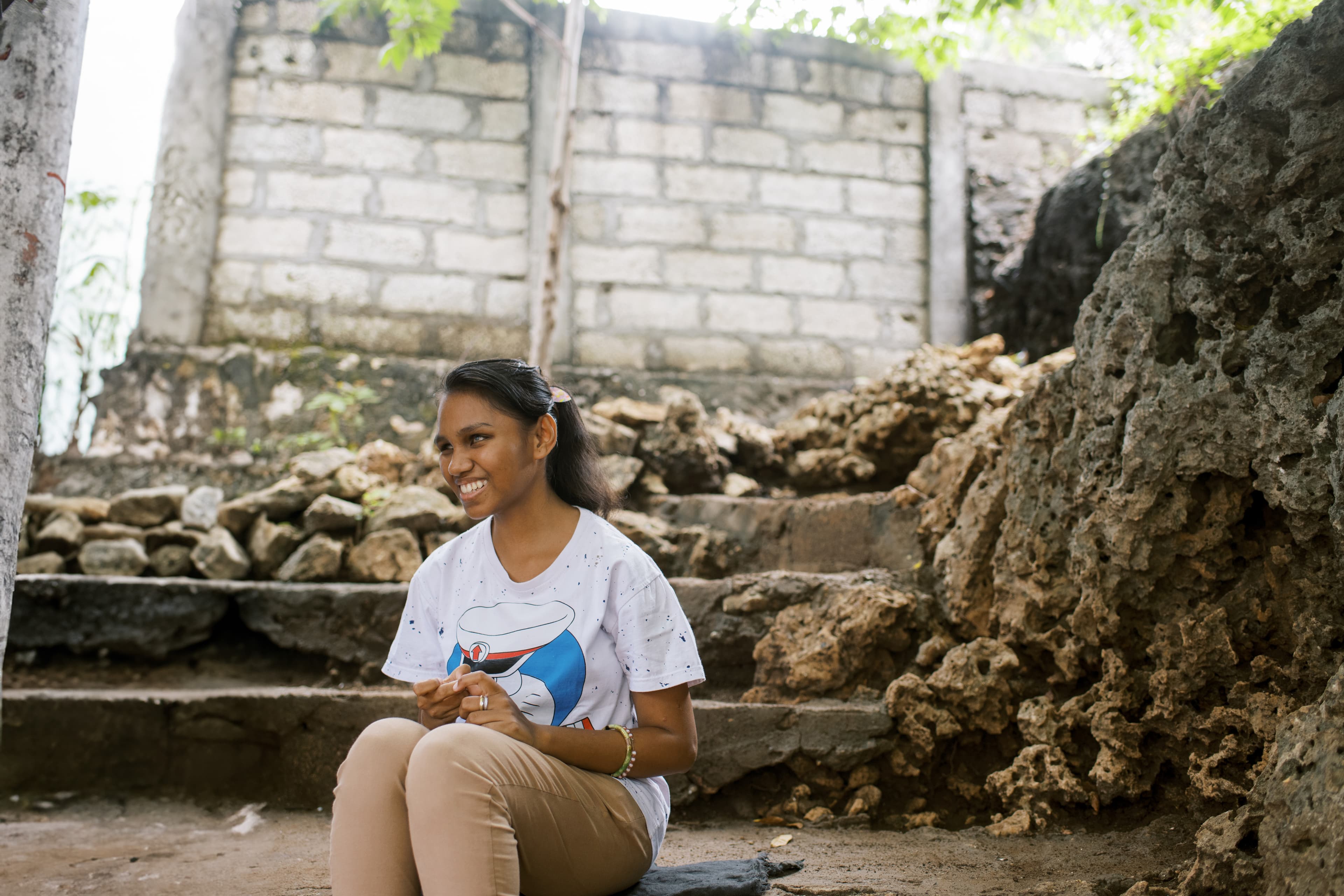 A young girl that faces blindness is sitting on the ground smiling with her hands in her lap.
