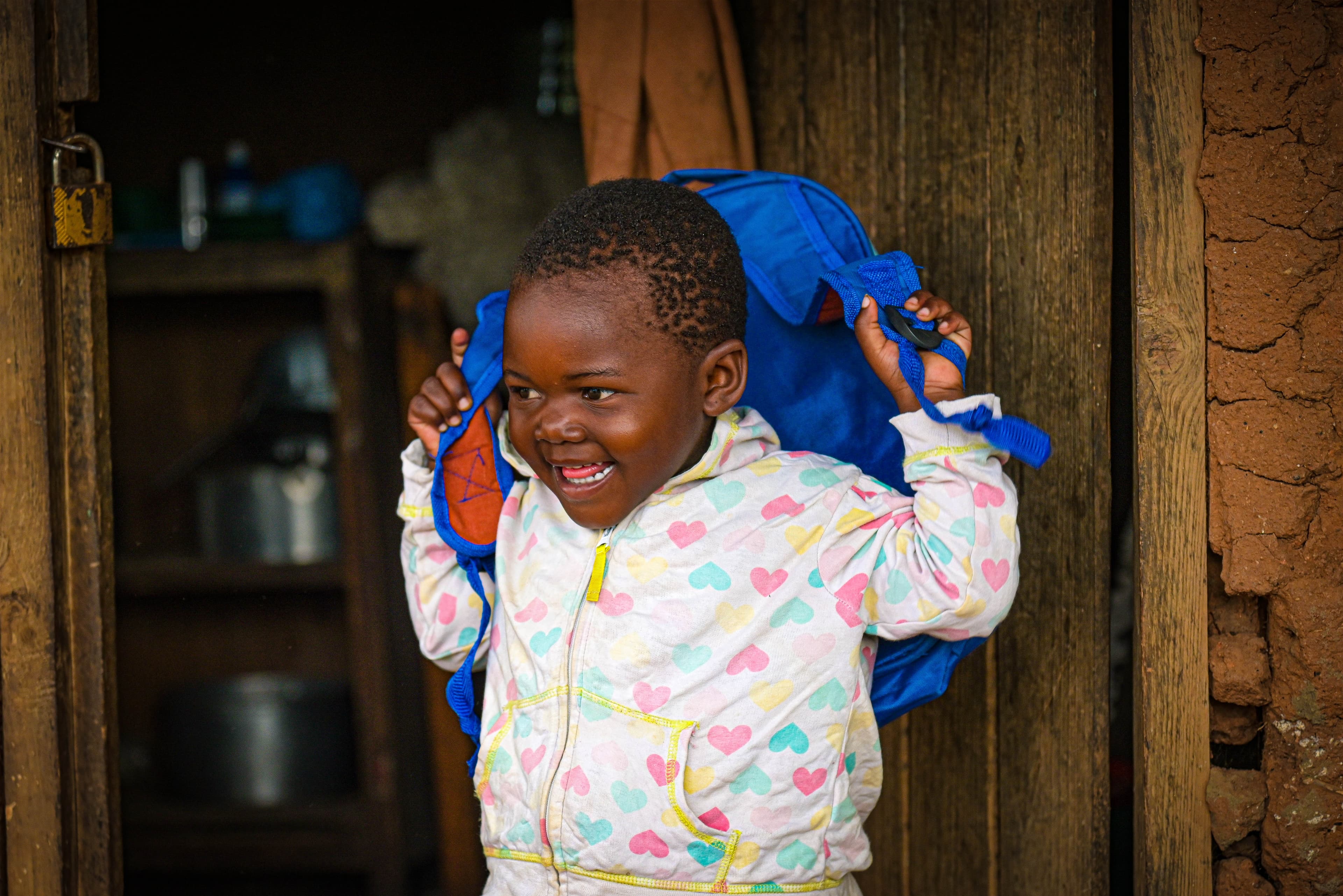 A young African girl holds a bright blue backpack behind her as she smiles brightly.