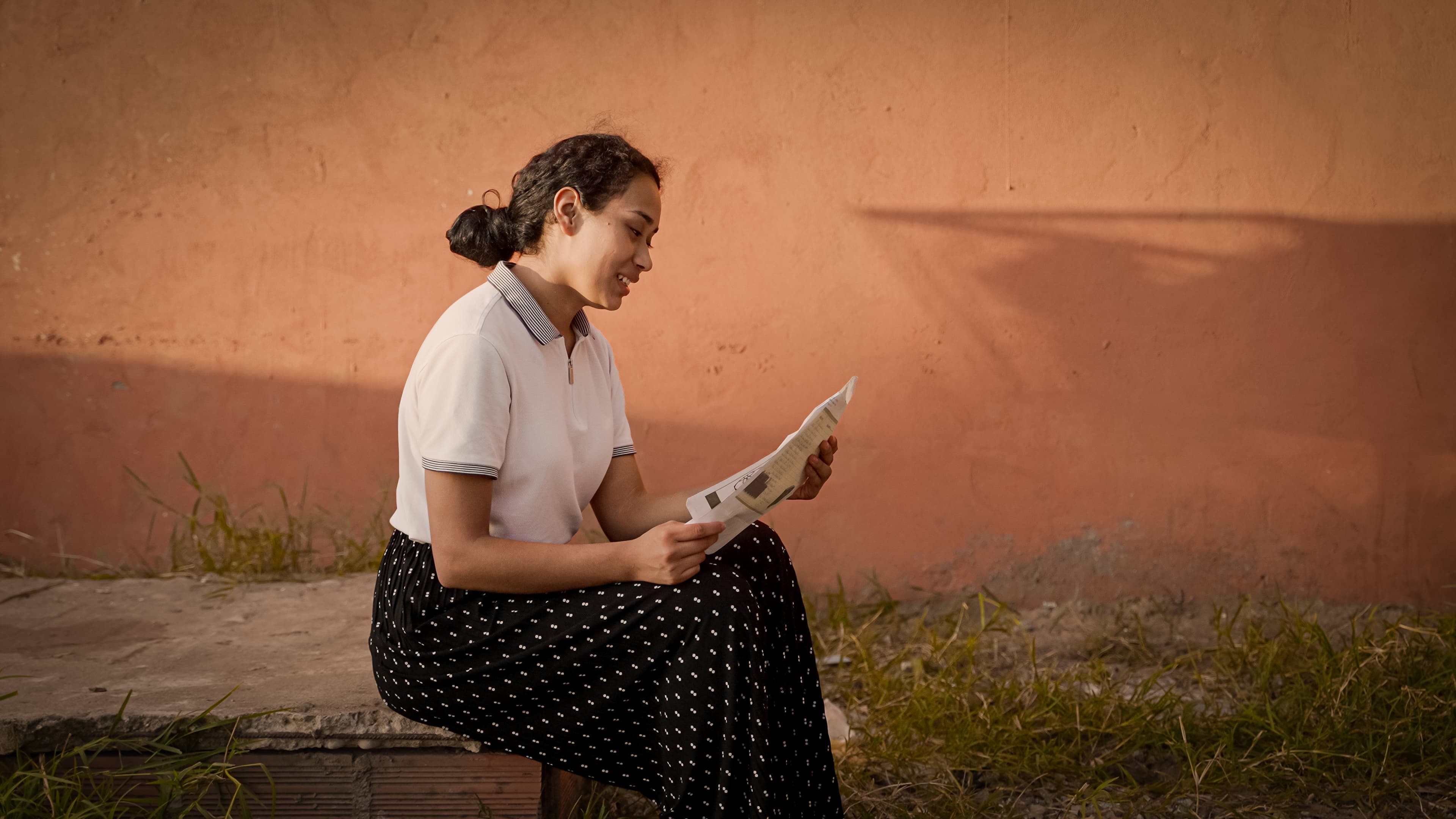 A teen Bolivian girl sits in front of an orange wall while reading a letter and smiling.