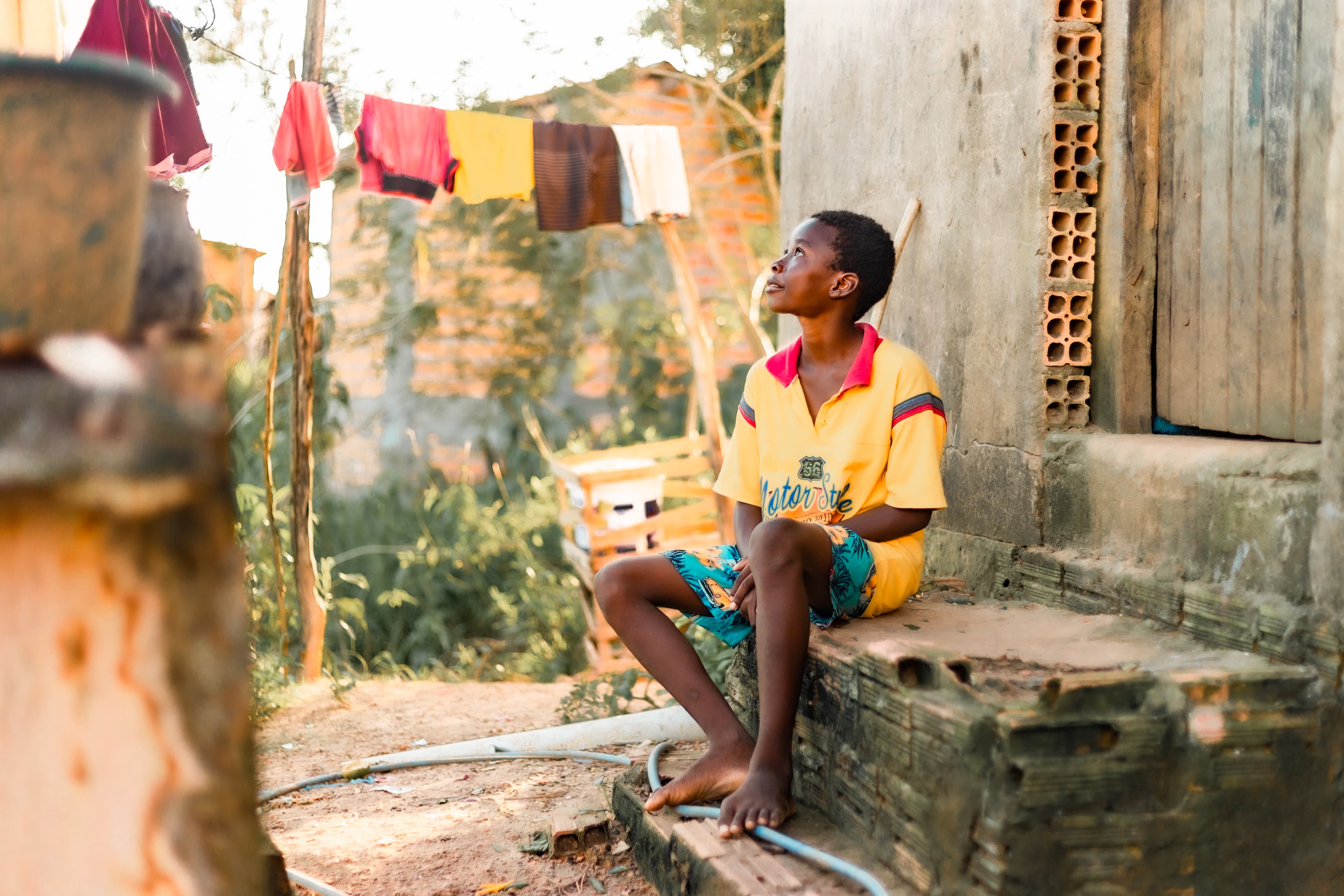 A boy is wearing a yellow shirt with blue patterned shorts. He is sitting on a step outside his home. In the background is a clothesline with clothes hanging on it.