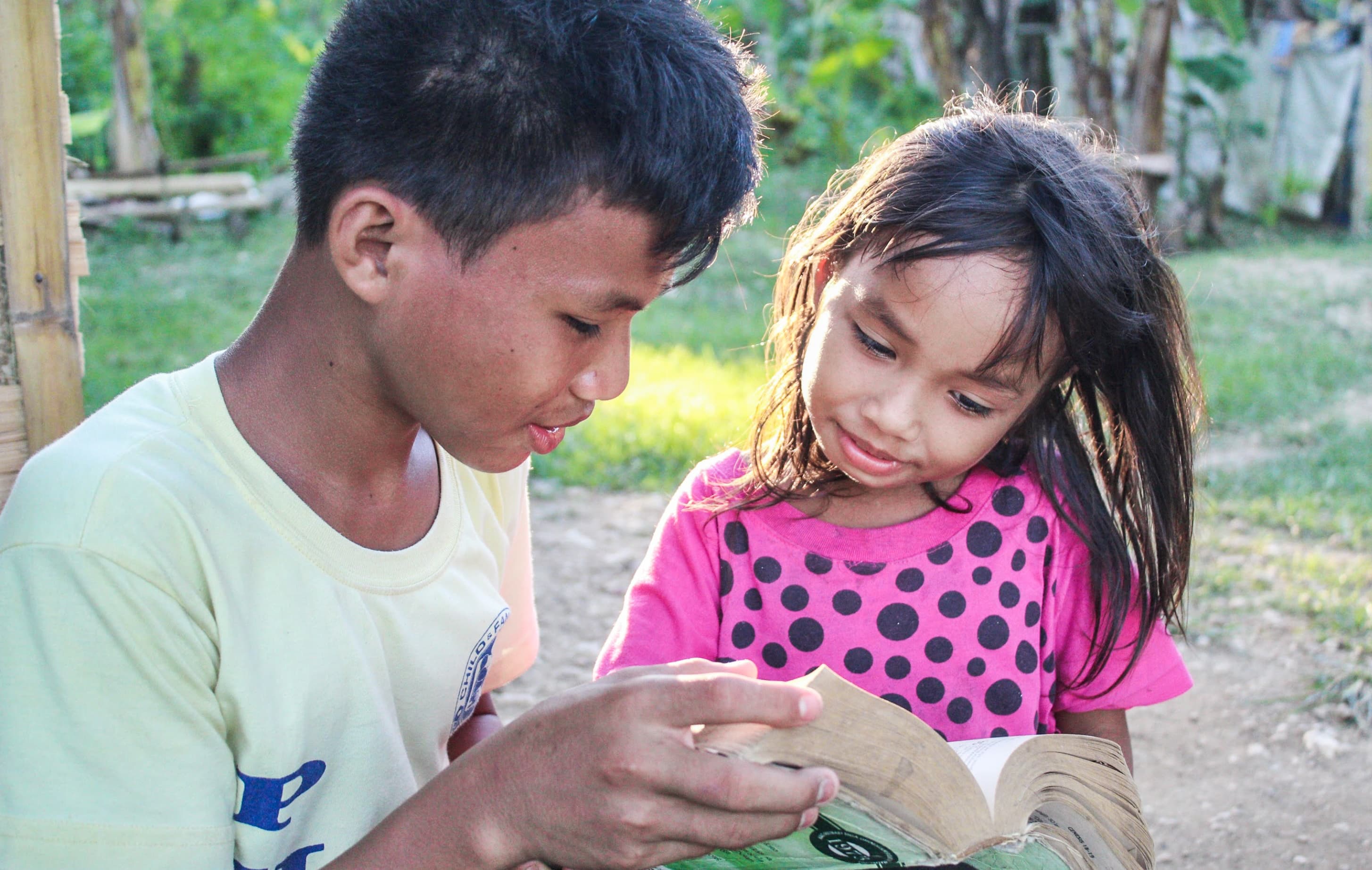 A boy wearing a yellow T-shirt reads from a Bible to a younger girl in a pink T-shirt.