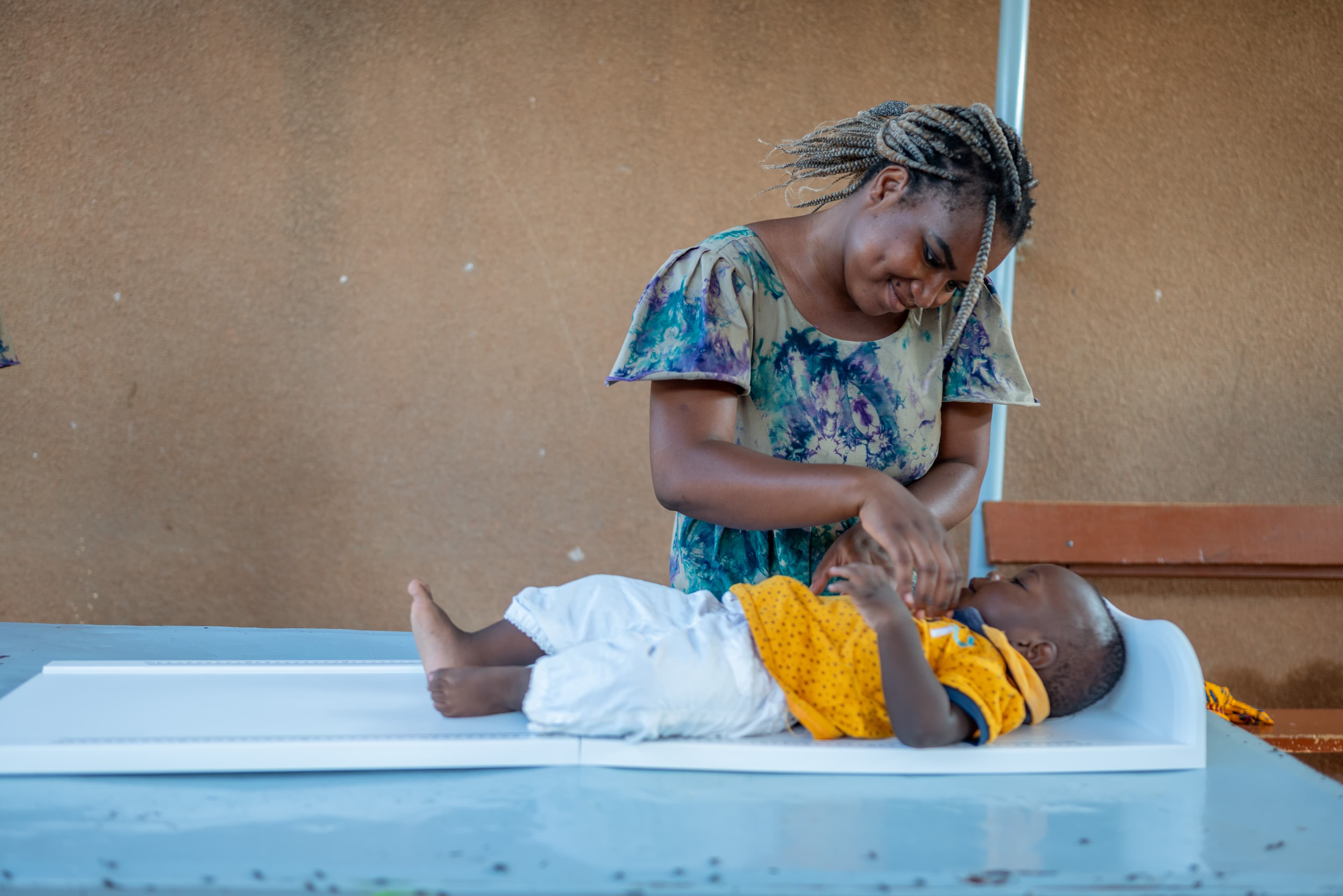 A mother wearing a blue dress stands behind her baby, who is wearing yellow and lying on a white table. The woman is smiling down at the baby.