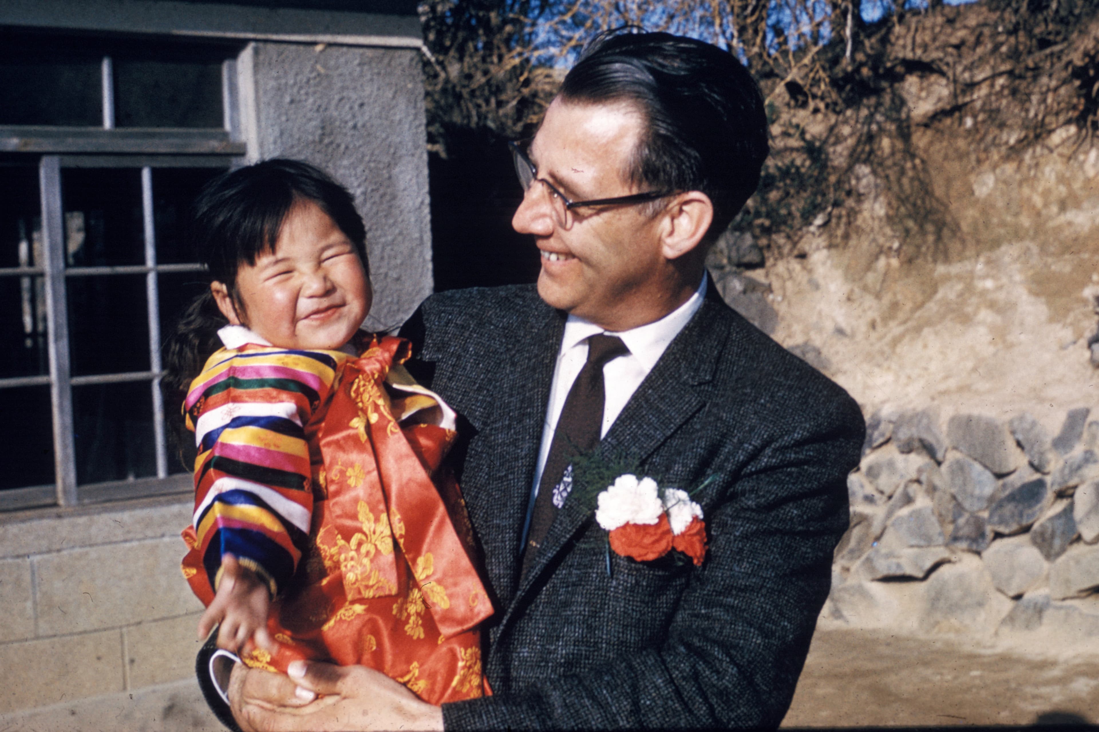 An older man wearing a tie smiles while holding a young Korean girl.