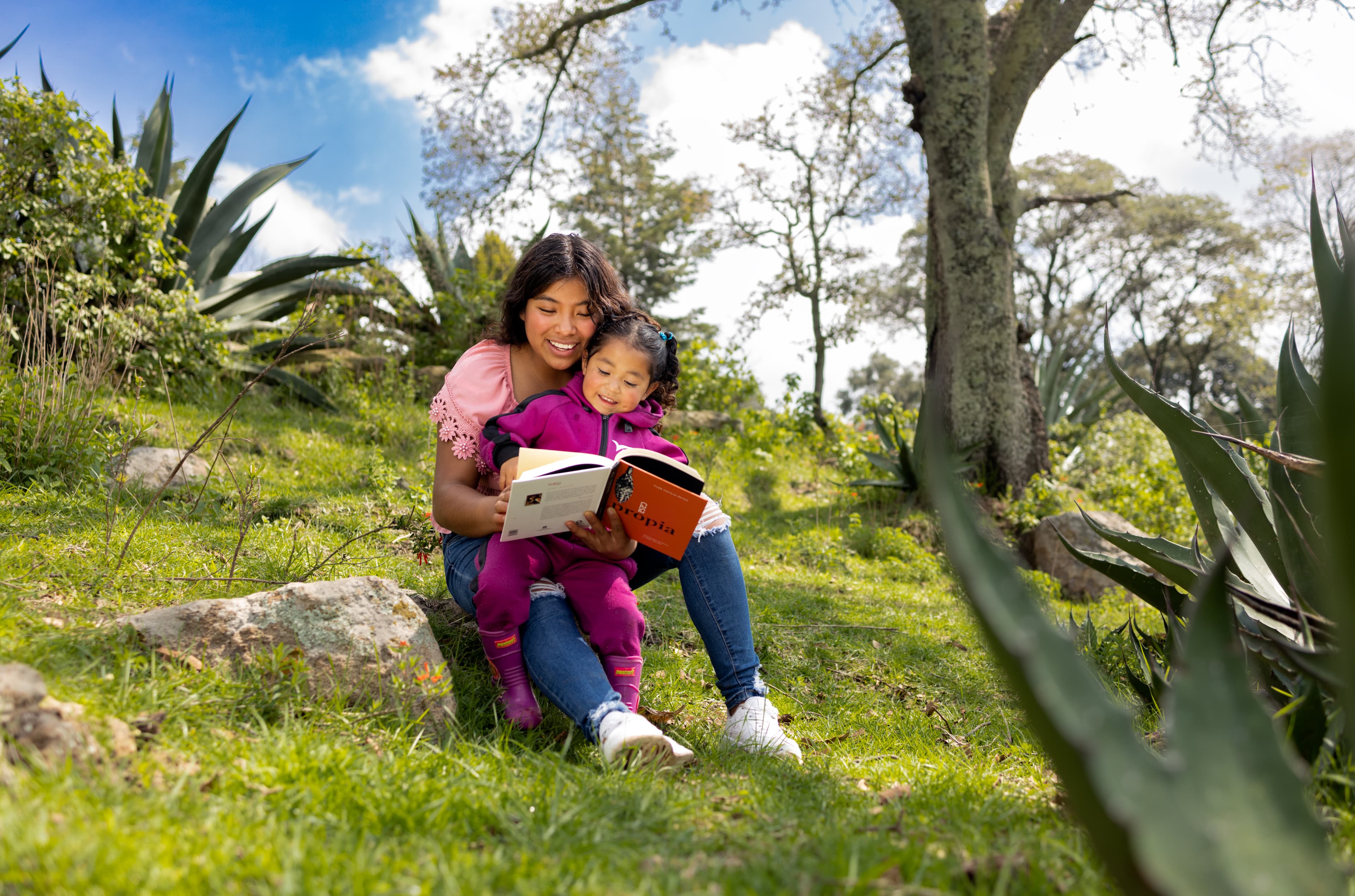 A mother and daughter read together while sitting on a grassy hill.