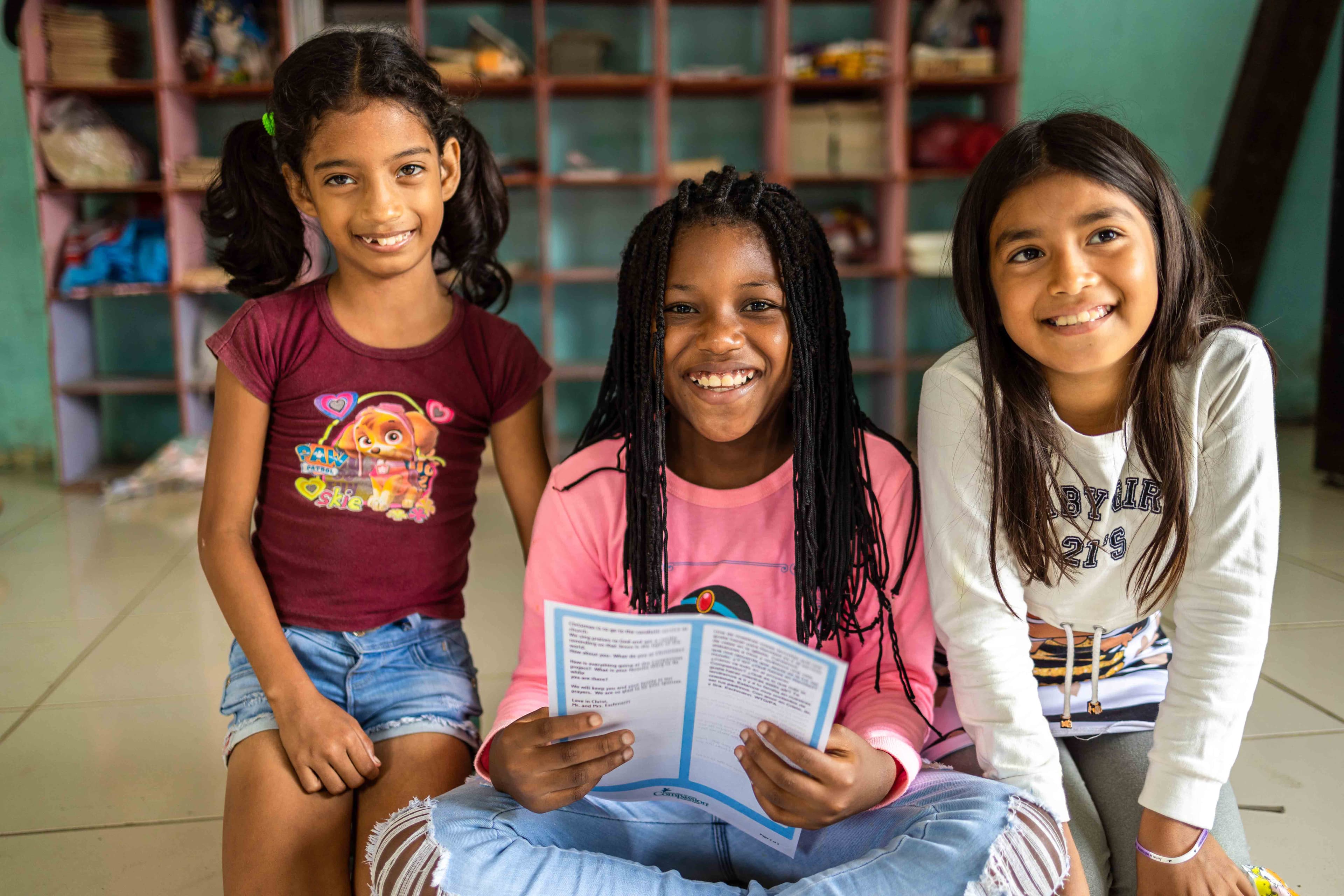 Three young girls are sitting and smiling as one holds a letter from her sponsor.