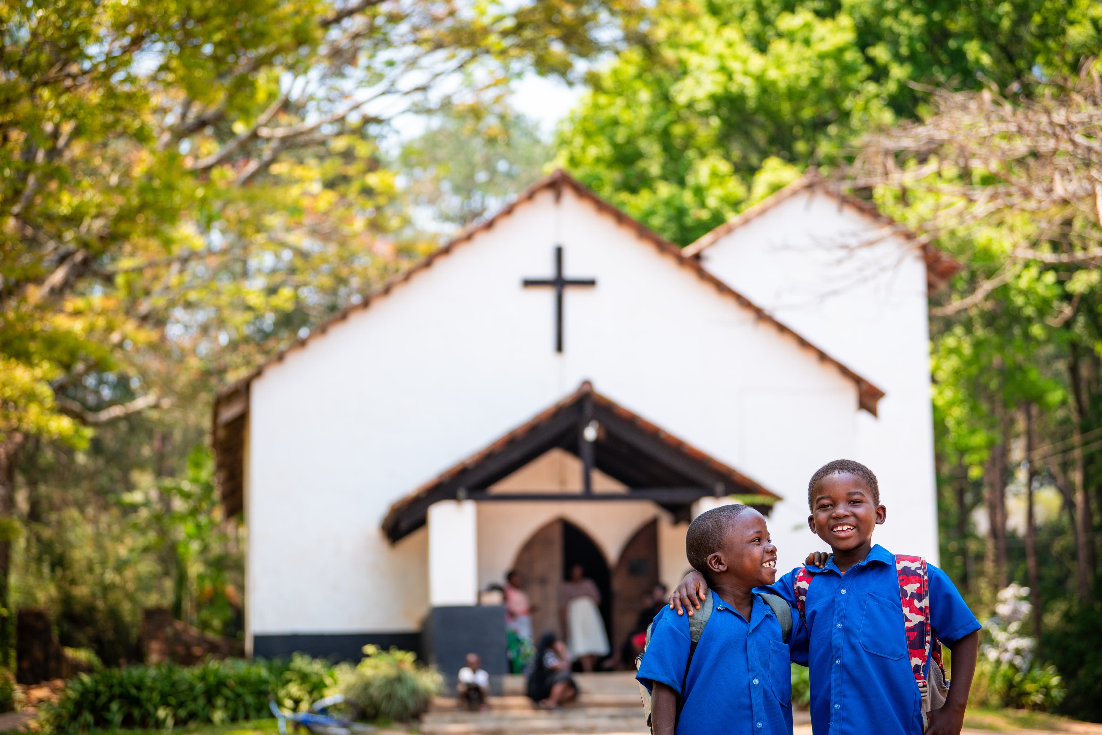 Two boys in blue shirts stand next to each other in front of a white church center building, and behind them a woman stands in the doorway of the church and others sit on the stairs going up to the building.