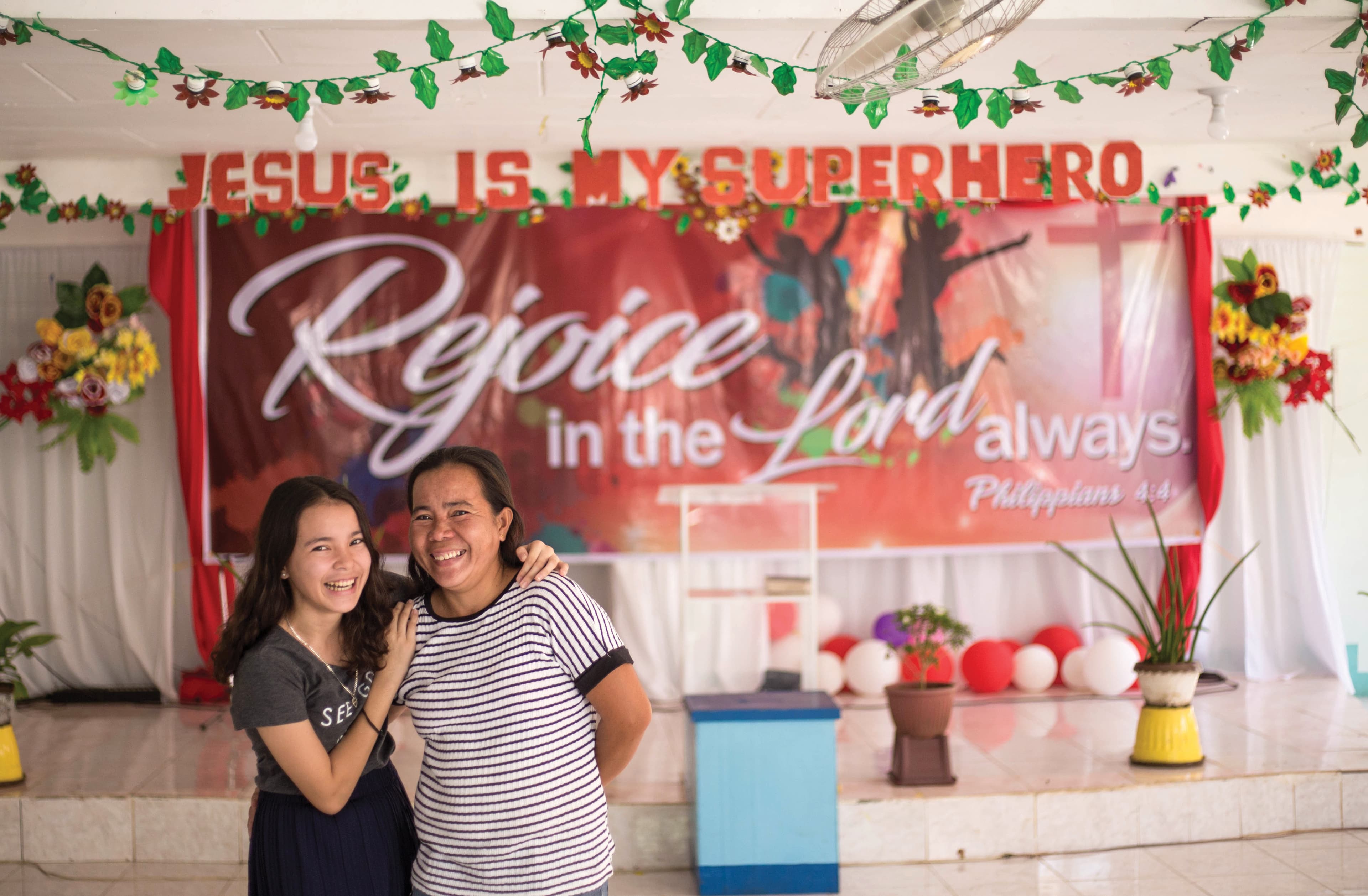Teenaged girl and older woman pose in front of banner that reads "Rejoice in the Lord always."