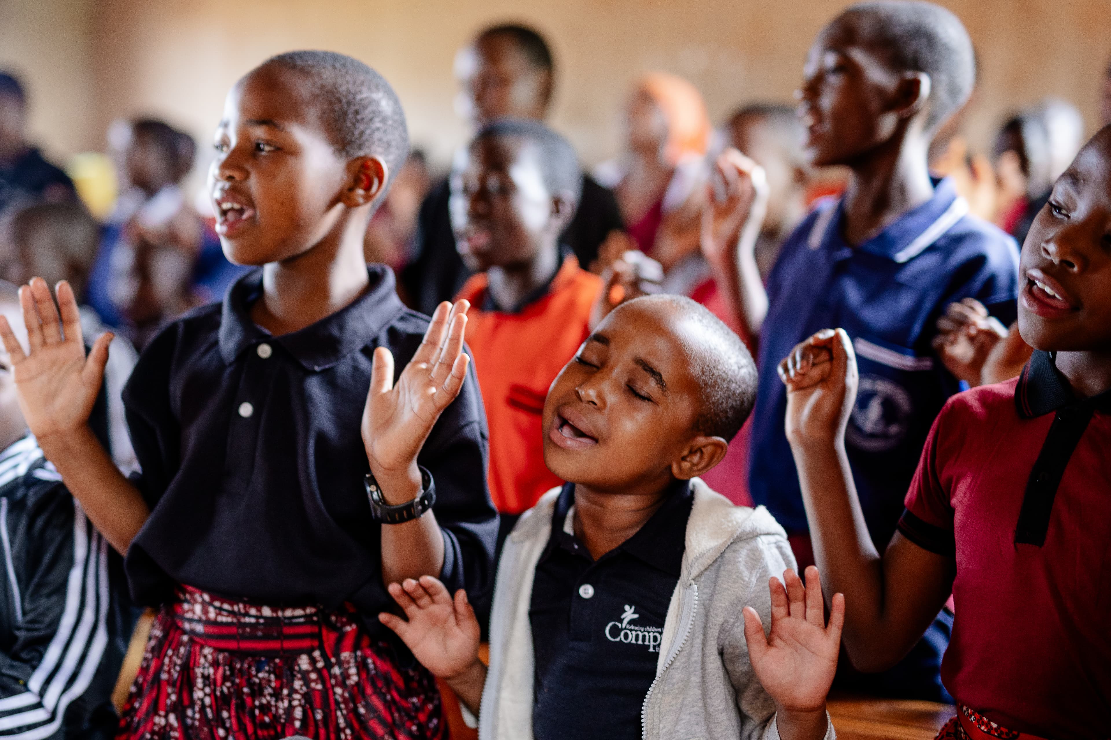 A young child holds a plate with bread and an egg on it, as well as a cup. He is smiling at the camera. Behind him is a line of children waiting to receive food from two cooks outside a Compassion center.