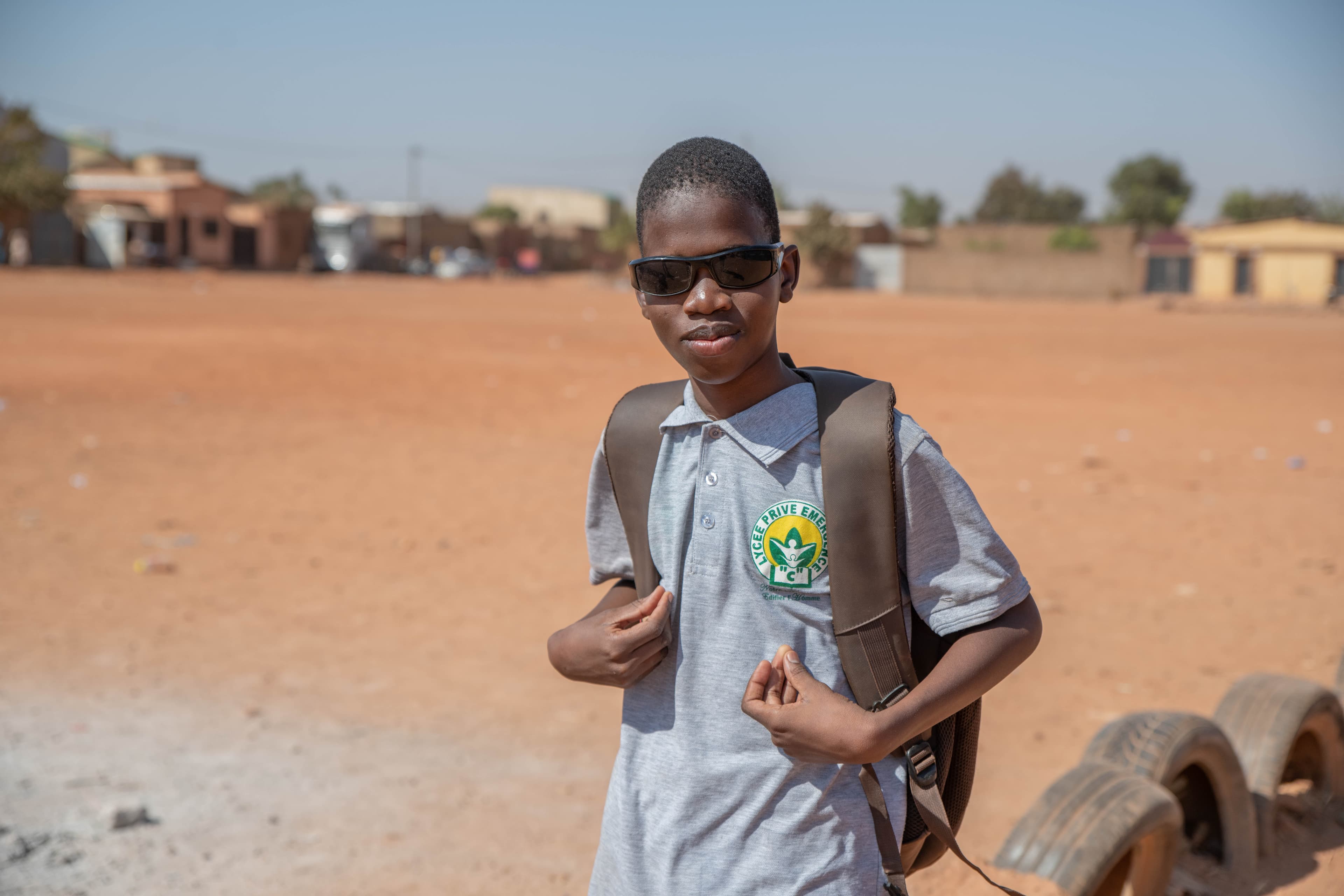 An African teen wears sunglasses and stands with a backpack on his back.