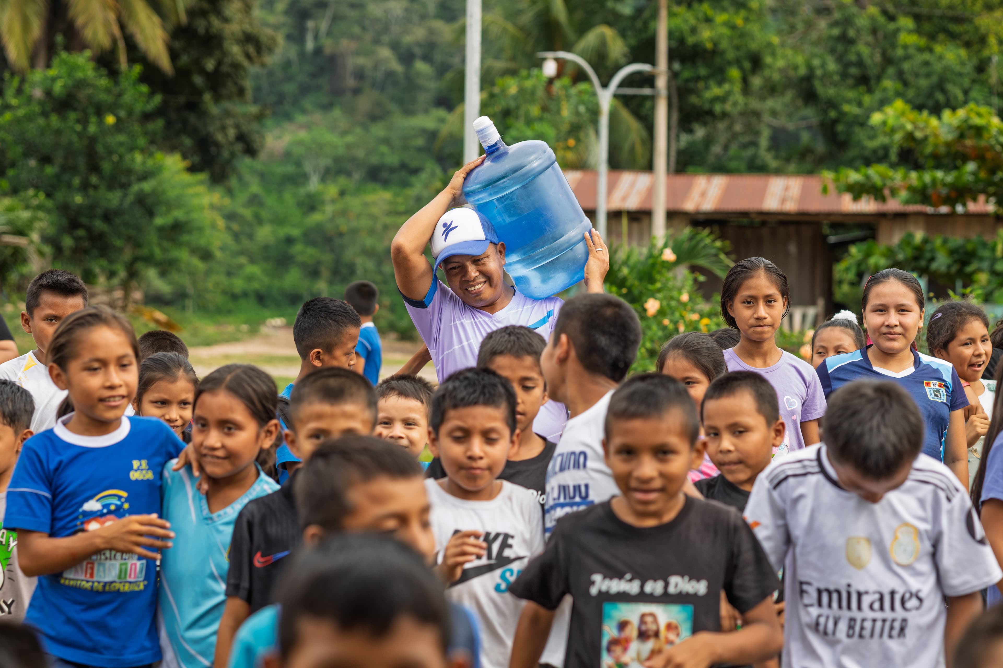 A group of children surround a man who is carrying a water container on his shoulder.