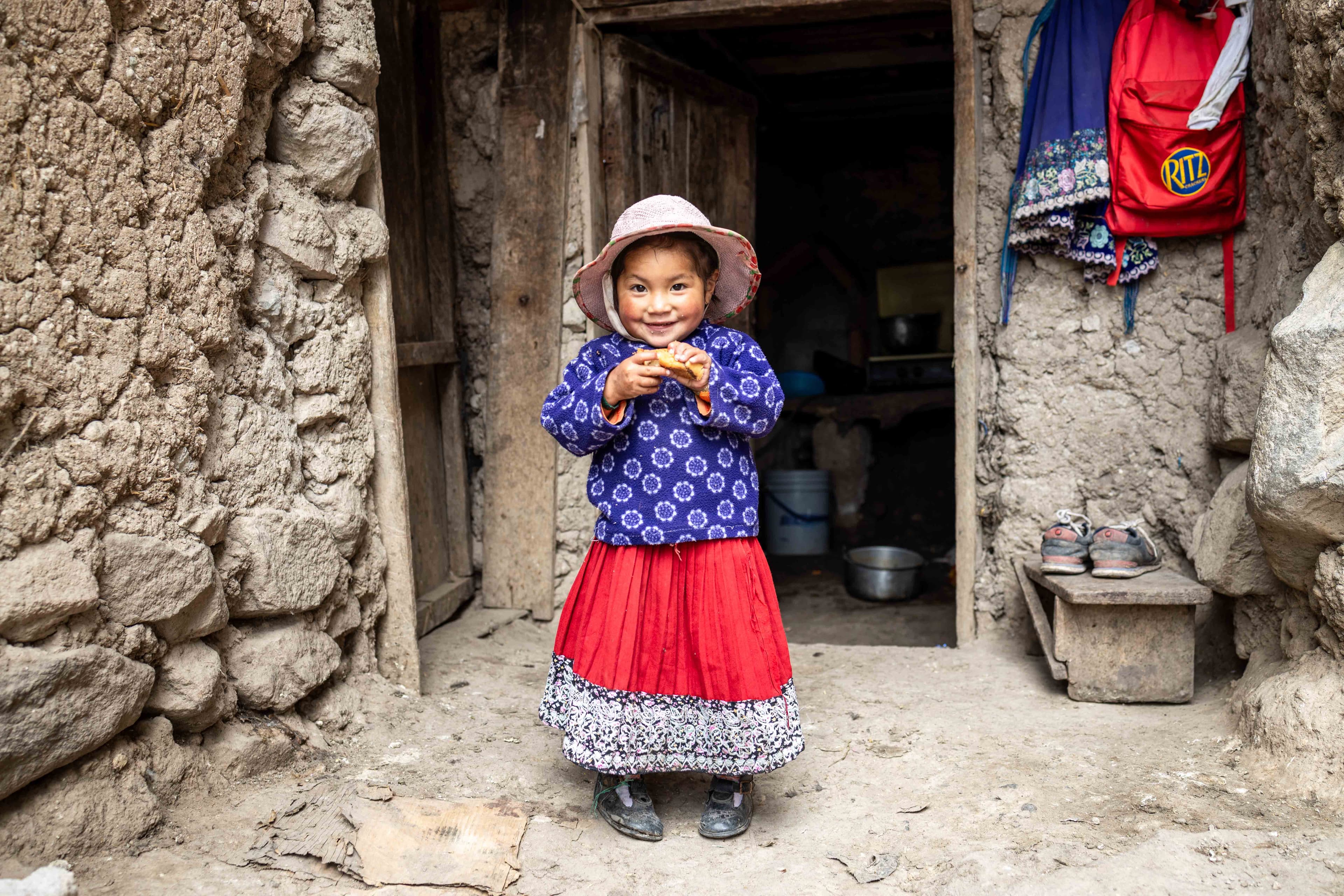 A young girl wearing a bright red skirt and hat smiles outside of her clay home.